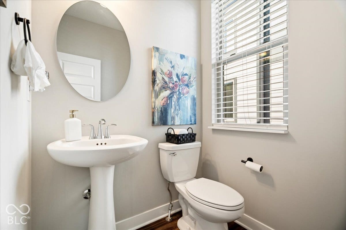 Pedestal sink with chrome faucet, oval mirror, toilet, window with blinds, and wood-finish flooring