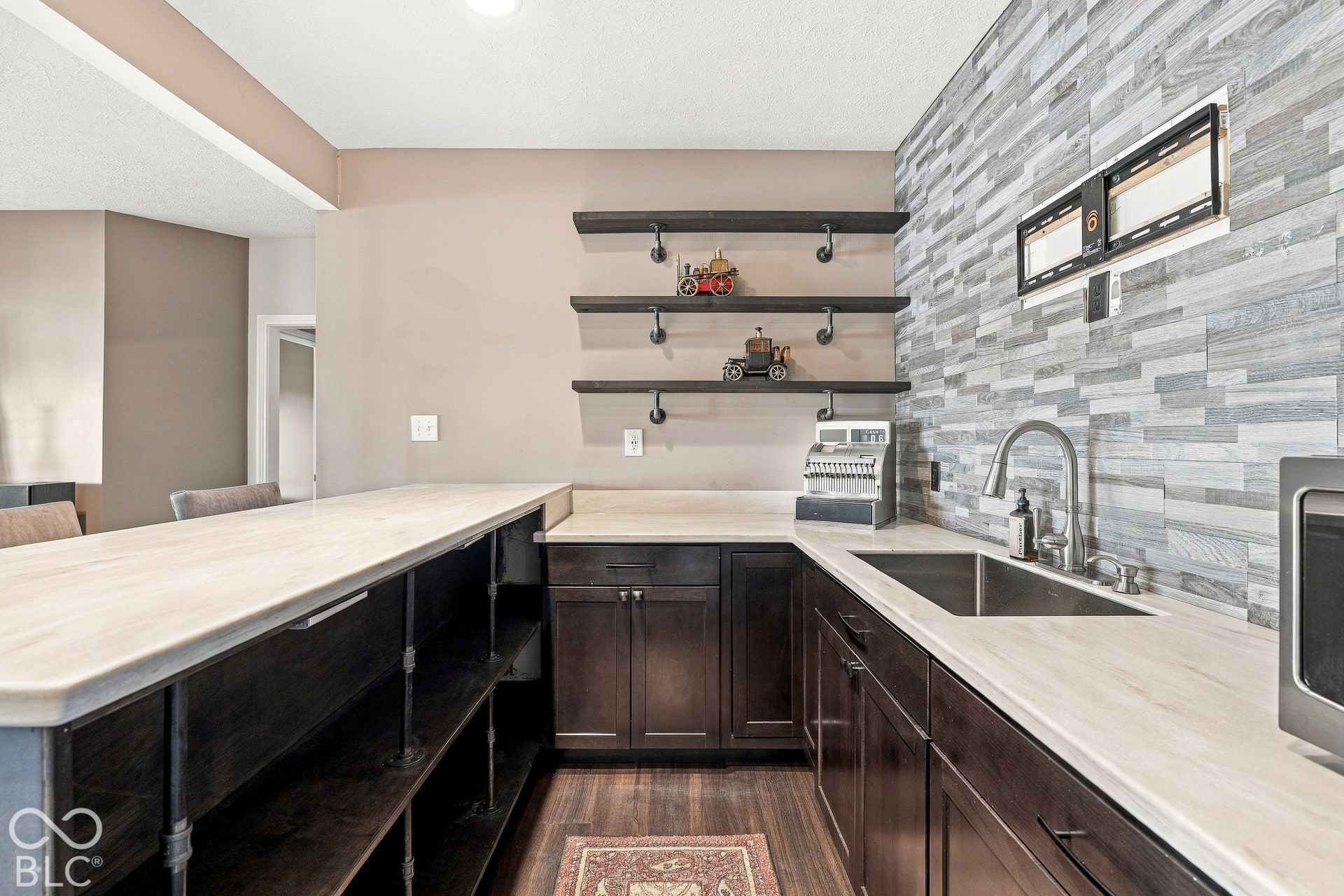 Custom bar area featuring dark wood-finish cabinetry, light-toned countertops, stone-tile backsplash, stainless steel under-mount sink with gooseneck faucet, and three-tier industrial pipe shelving