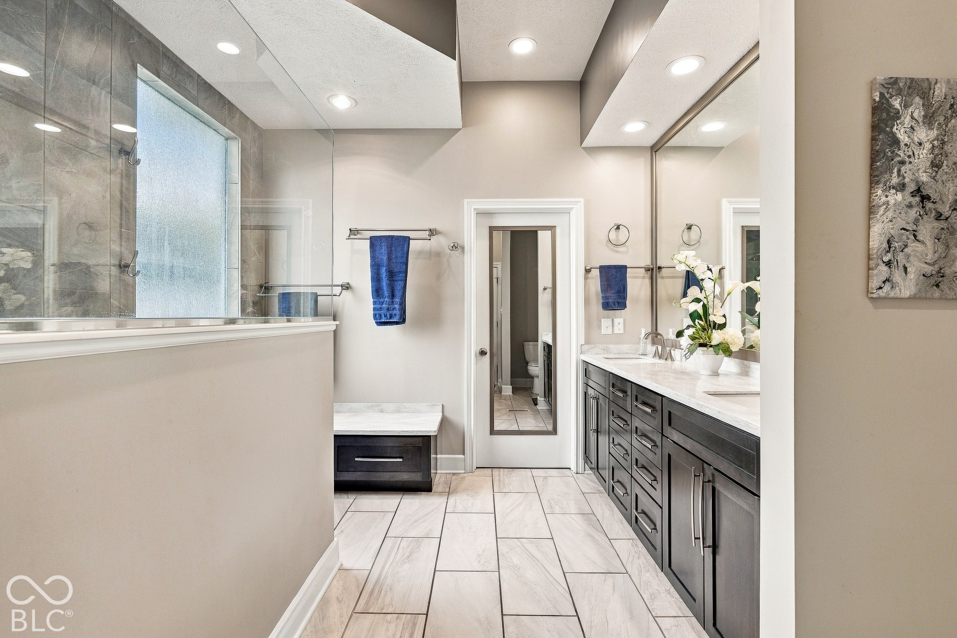 Bathroom featuring dark-finish cabinetry with a stone countertop, dual undermount sinks, frameless glass-enclosed shower with tiled surround, and rectified porcelain floor tile