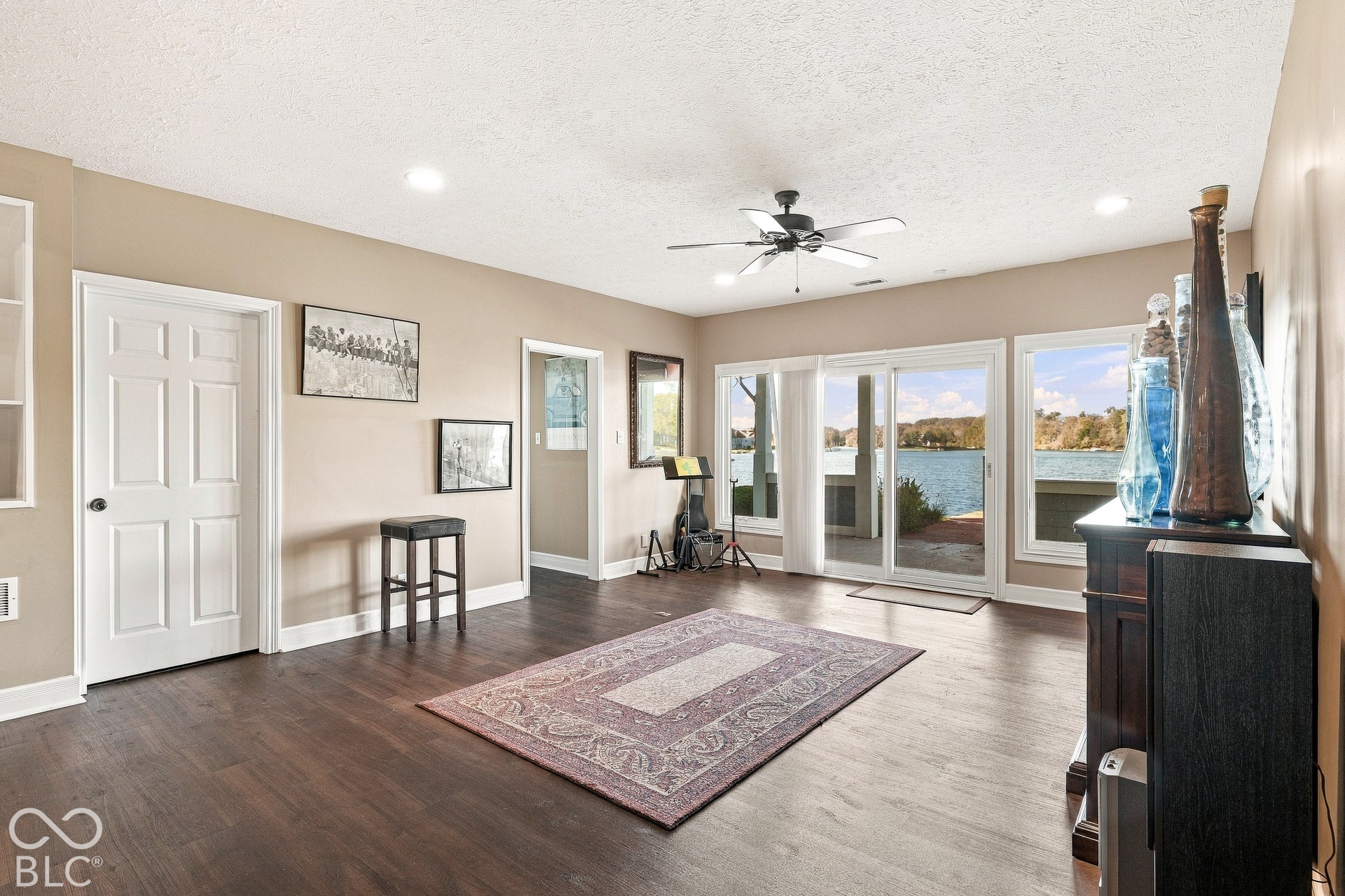 Spacious great room featuring multiple glass sliders opening to waterfront vistas, dark wood-finish flooring throughout, recessed ceiling lighting, and ceiling fan