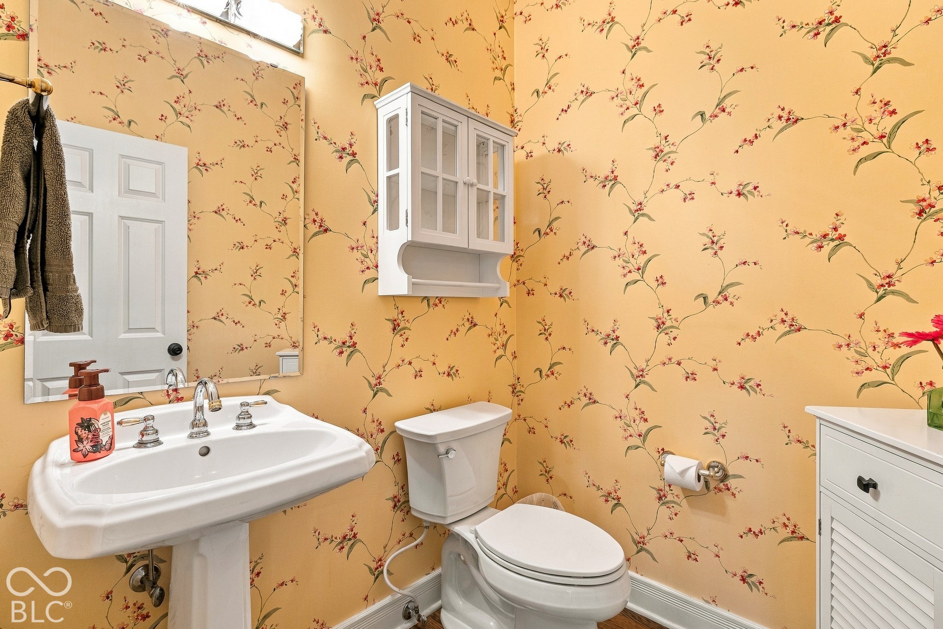 Ceramic pedestal sink with polished chrome widespread faucet, wall-mounted cabinetry with glass-front doors, and floral-patterned wallpaper