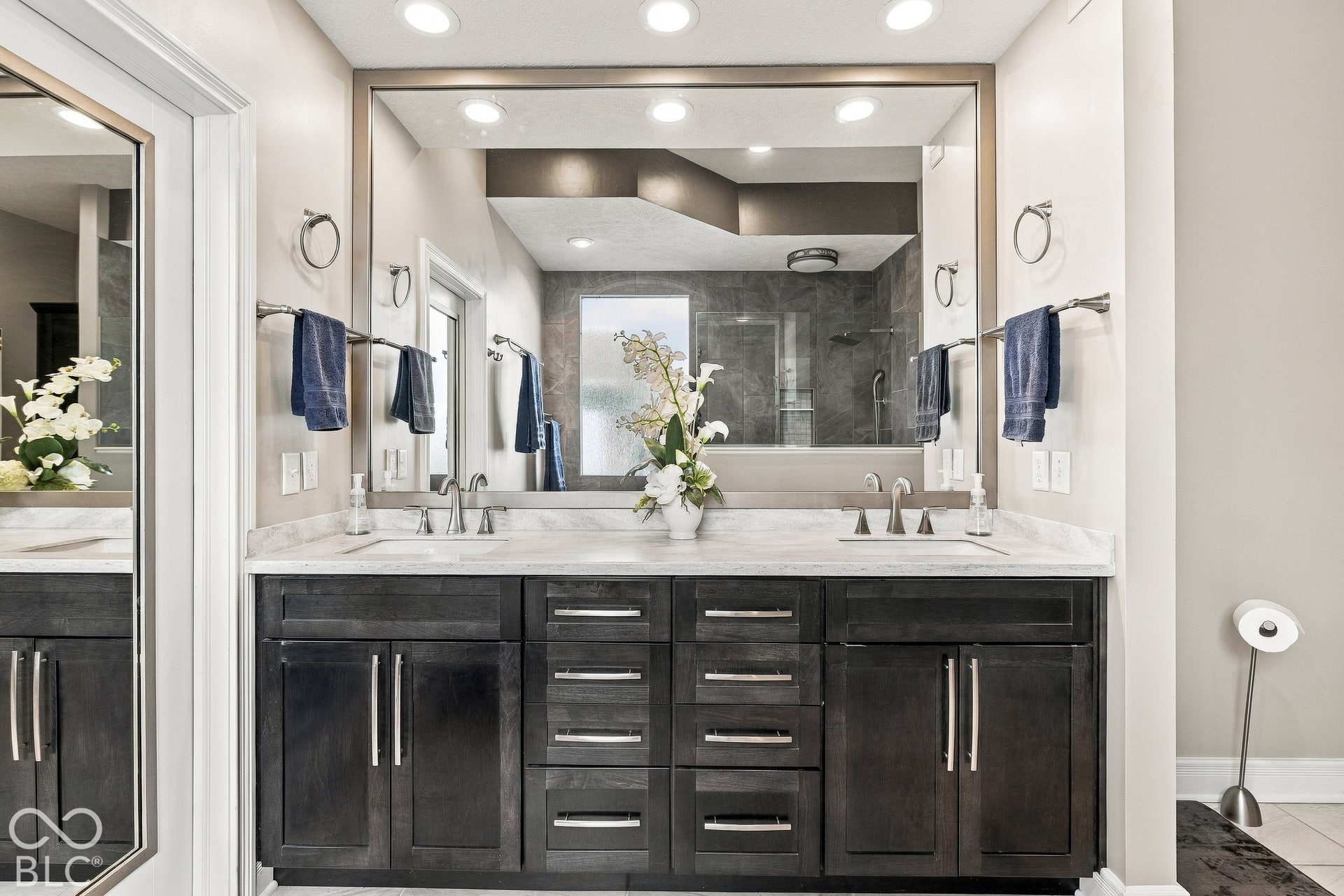 Dual-sink vanity featuring dark-toned cabinetry, light-toned stone-finish countertop, brushed-metal hardware, and a large framed mirror with recessed lighting