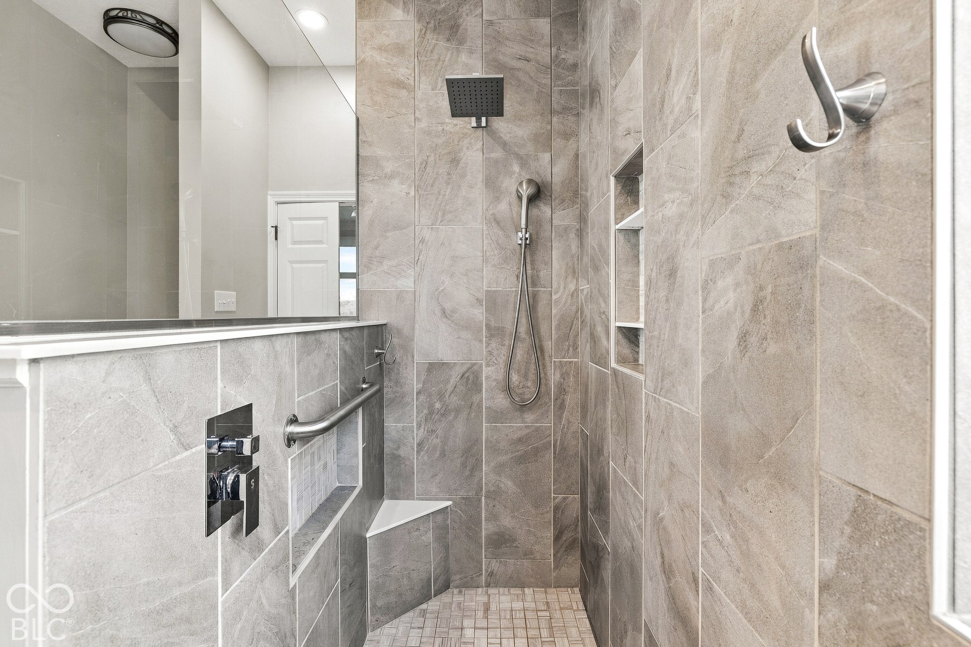 Walk-in shower featuring floor-to-ceiling stone-finish tile, rainfall showerhead, integrated tiled bench, recessed shelving, and a brushed metal grab bar