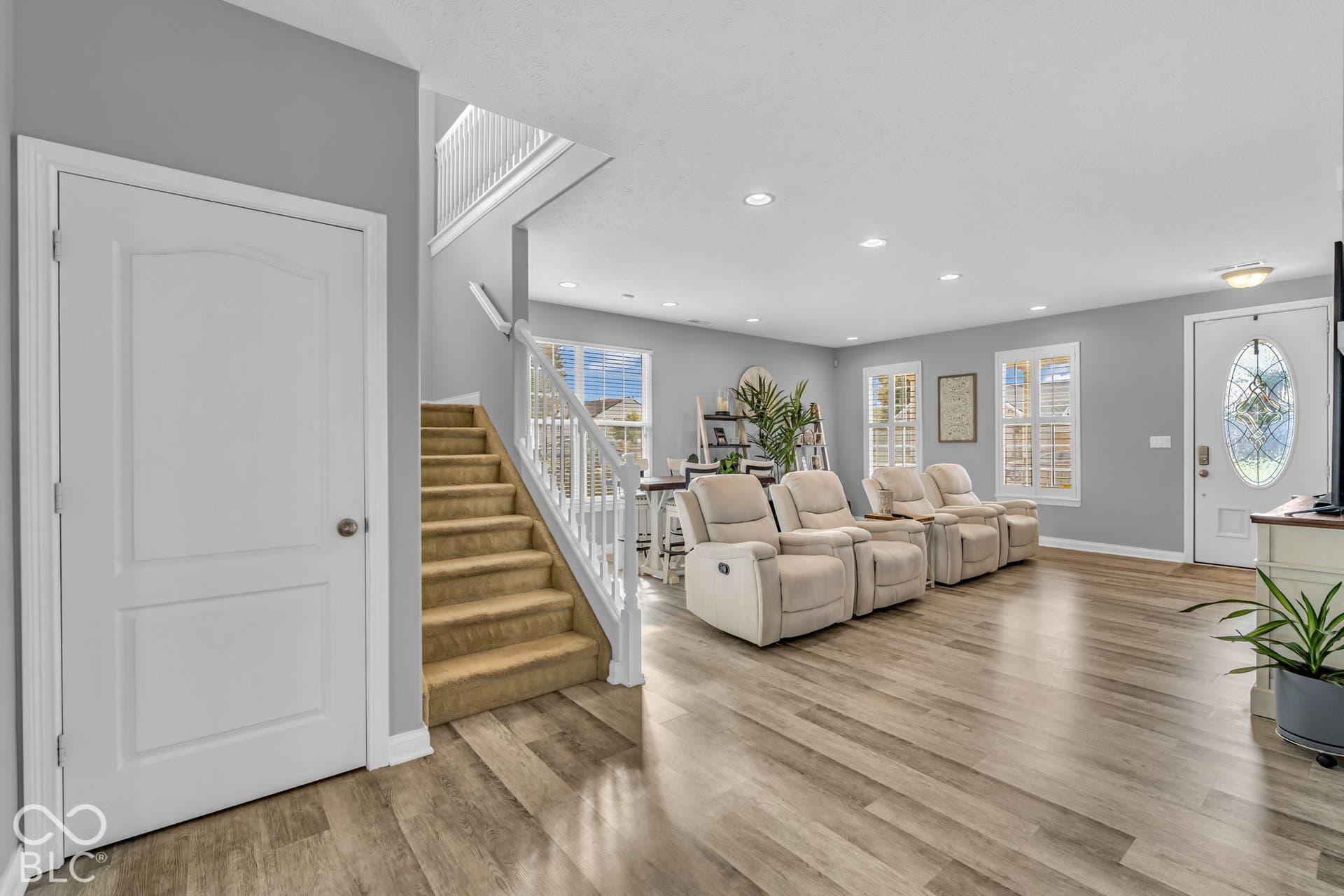 Expansive living area with wood-finish flooring, recessed lighting, multiple windows, and a carpeted staircase with white railings