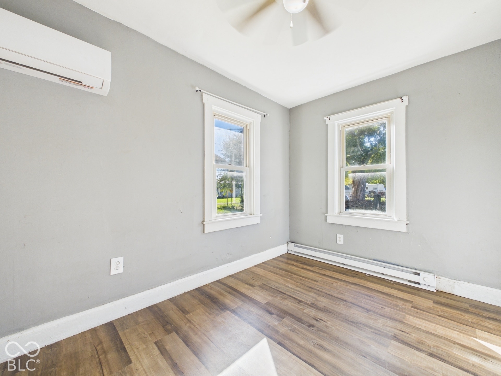 Wood-finish flooring, two windows with white trim, ceiling fan, and wall-mounted air conditioning unit