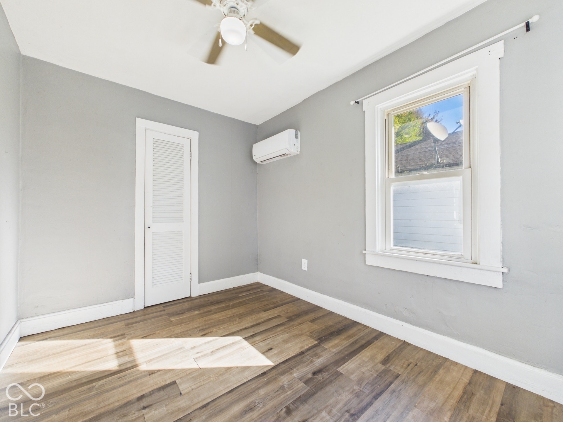 Bright corner bedroom featuring wood-finish flooring, neutral gray walls, a ceiling fan with integrated lighting, large double-hung window with natural light, and a louvered closet door for convenient storage access