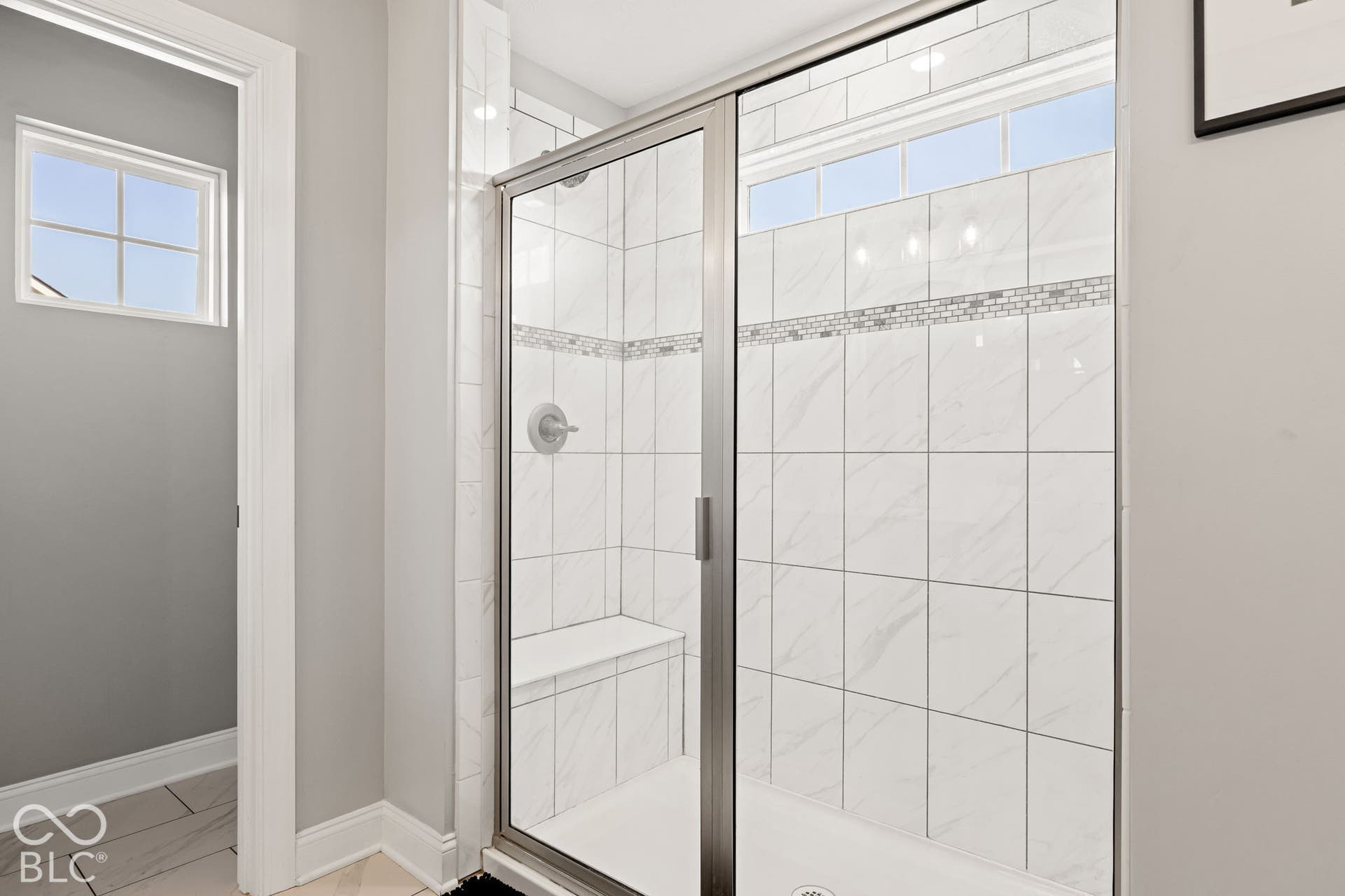 Walk-in shower with clear glass enclosure, white tile surround, mosaic accent band, dual shower heads,  two built-in benches, and brushed nickel fixtures