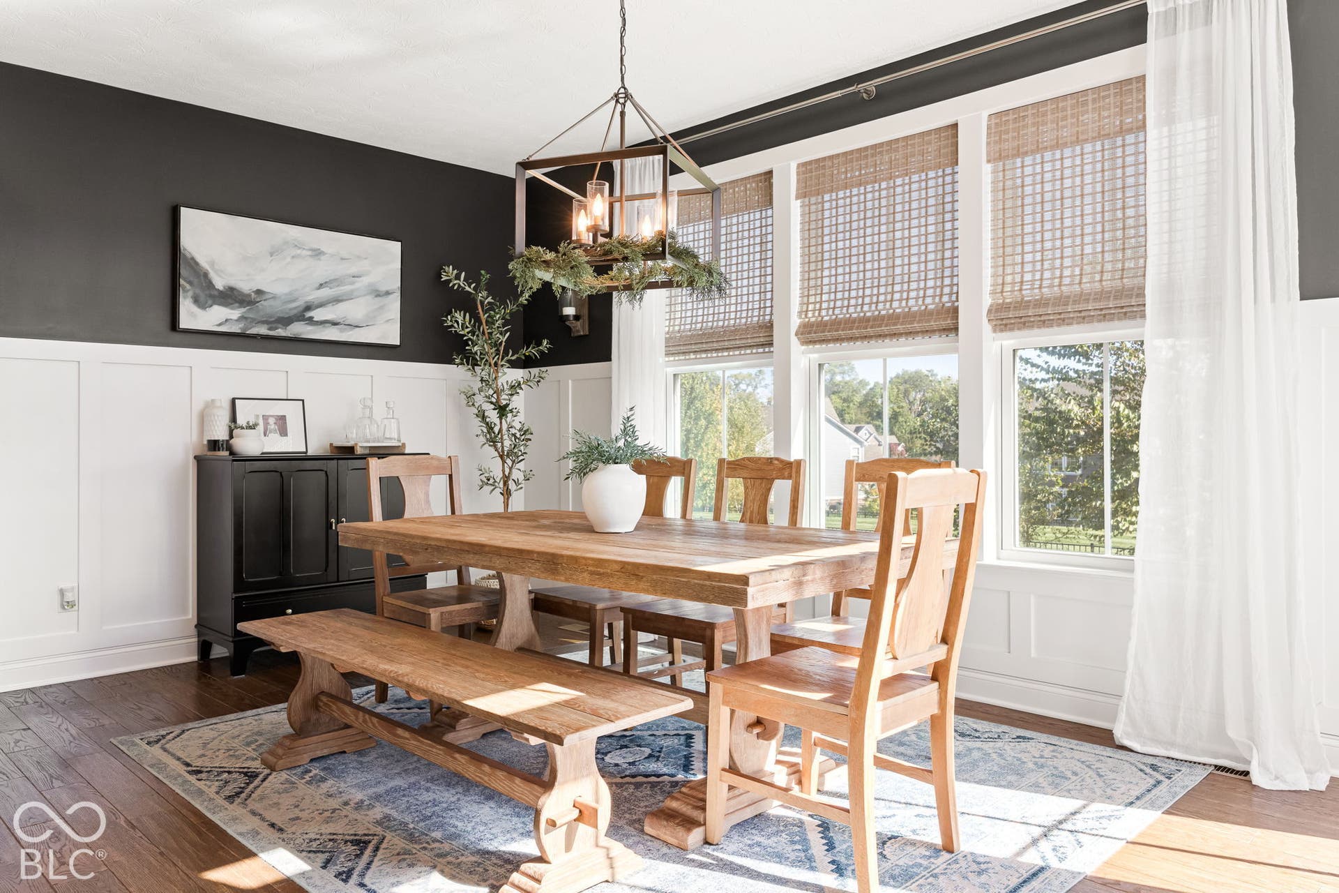 Hardwood flooring, wainscoting, multiple windows with woven Roman shades, and a large central chandelier
