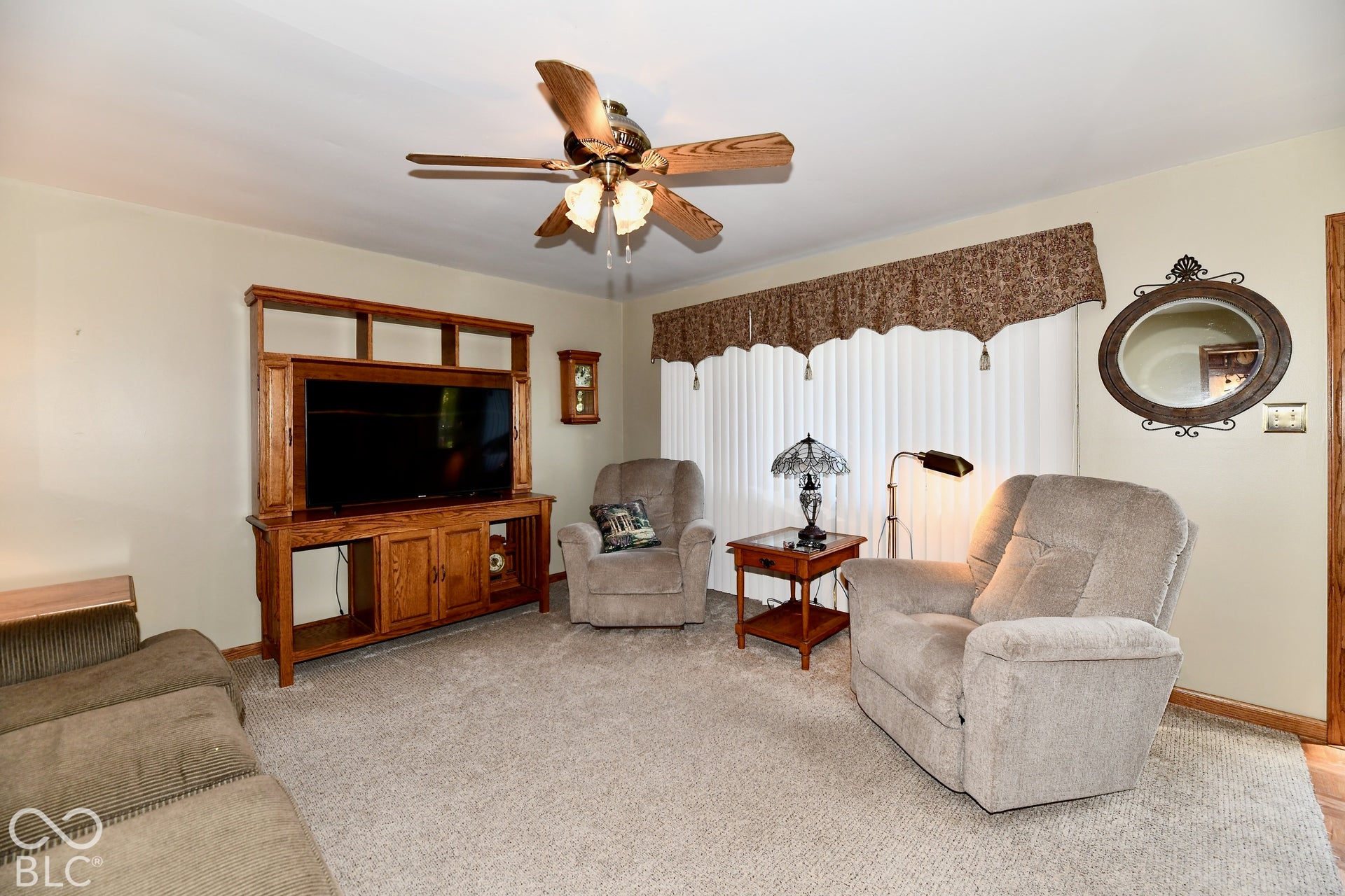 Spacious living room featuring a ceiling fan with light fixture, decorative window treatments with valance, and neutral carpet flooring