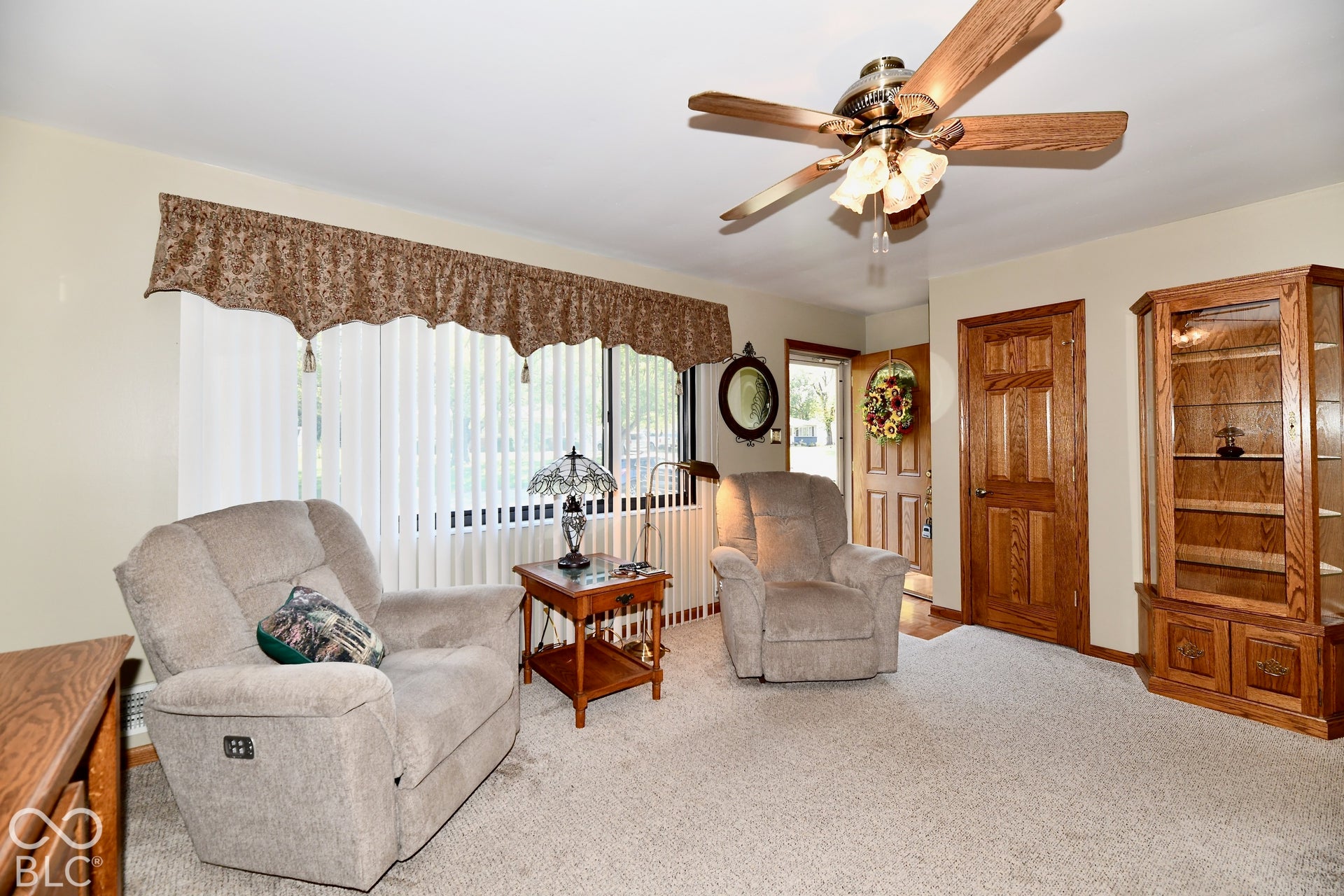 Carpeted flooring, wood-finish ceiling fan, wood-finish entry door, and large window with vertical blinds
