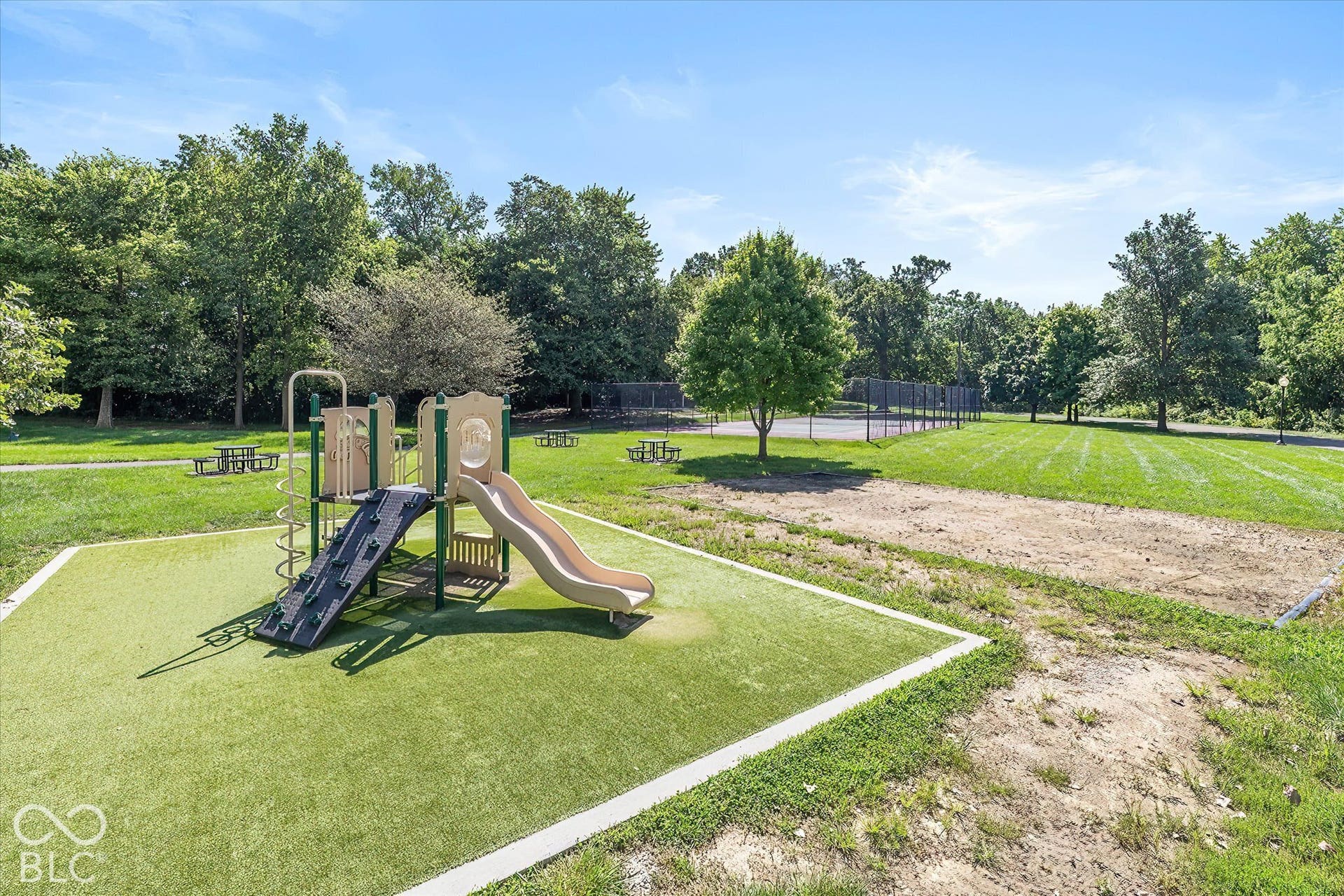 Community Playground:  Recreational area featuring a playground with artificial turf surfacing, a paved pathway, a tennis court with chain-link fencing, and picnic tables