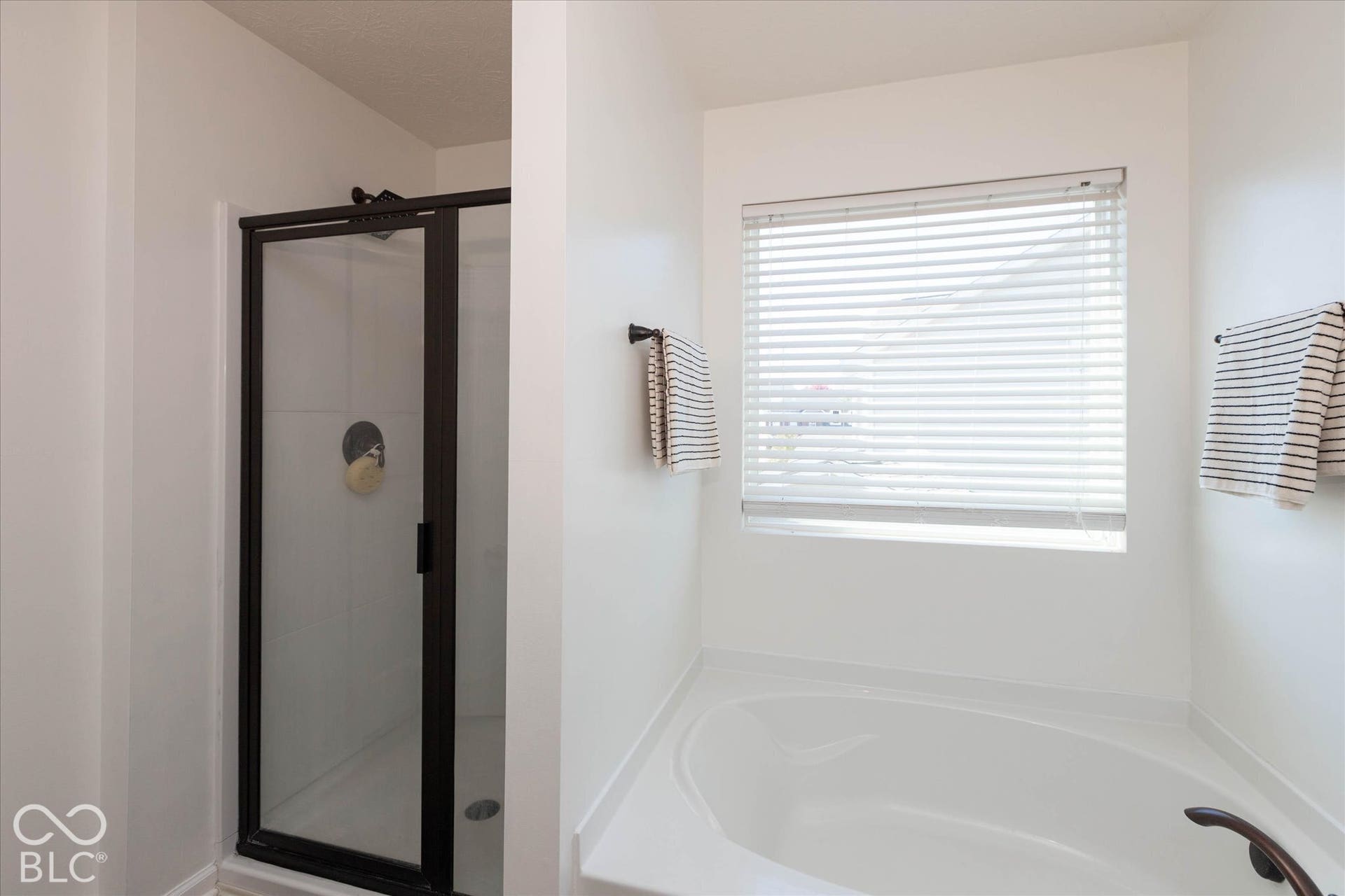 Enclosed shower with dark-framed glass door, white soaking tub, and window with horizontal blinds