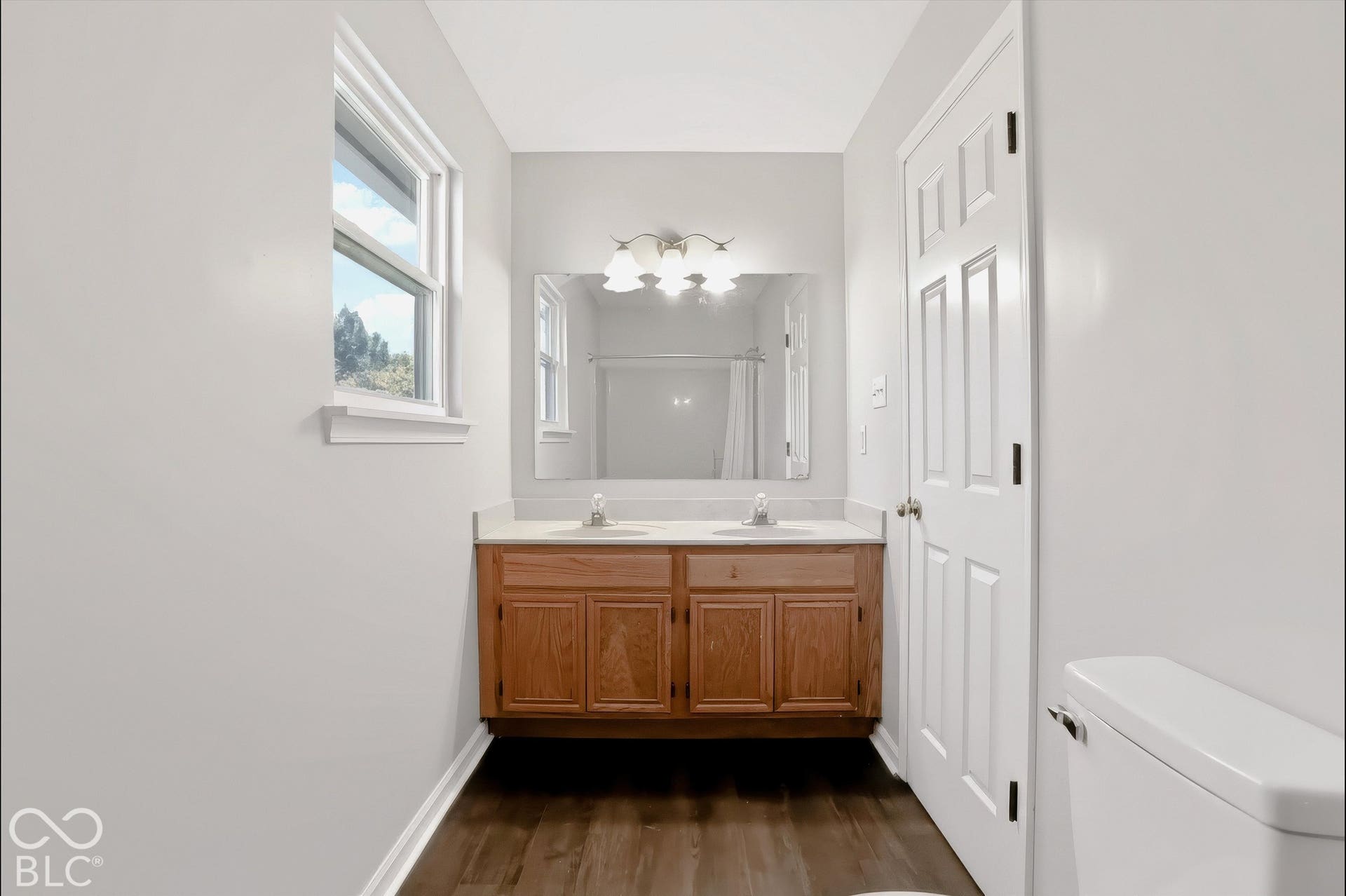Virtually Altered - Dual vanity with wood-finish cabinetry and light-toned countertop, framed mirror, and wood-finish flooring