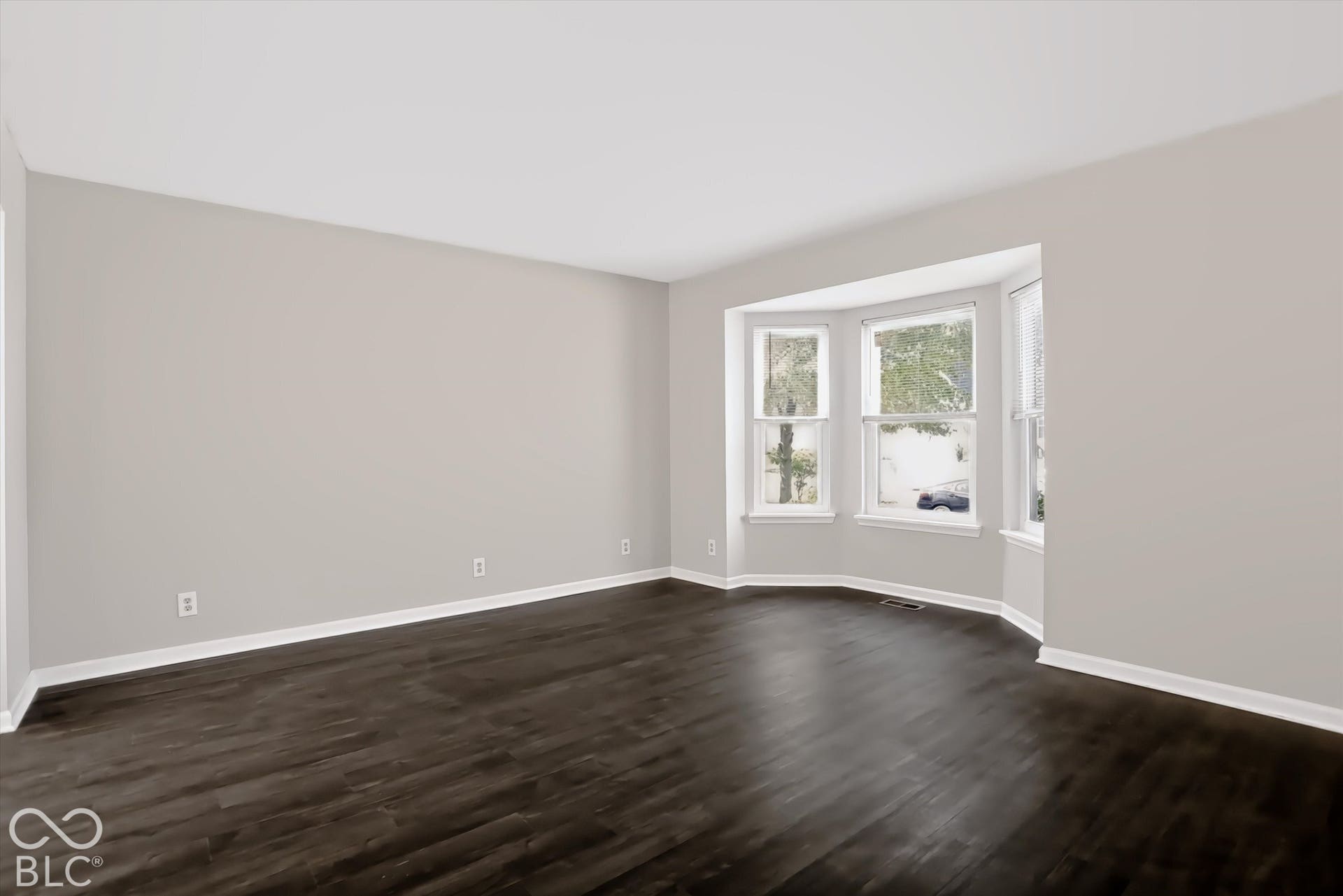 Expansive room with dark wood-finish flooring, light neutral wall paint, and a prominent bay window feature