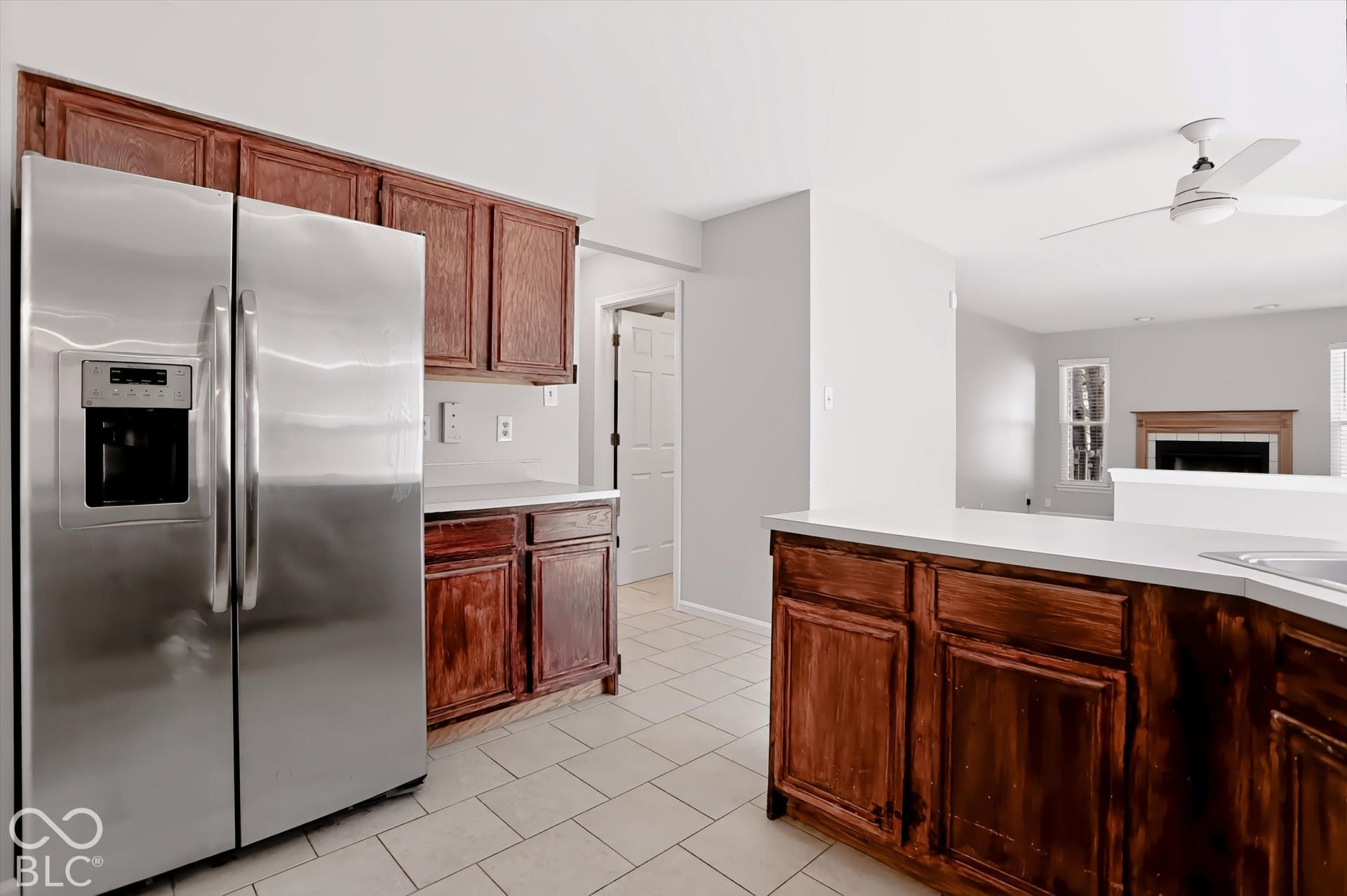 Stainless steel side-by-side refrigerator, wood-finish cabinetry, white countertops, tile flooring, and a ceiling fan