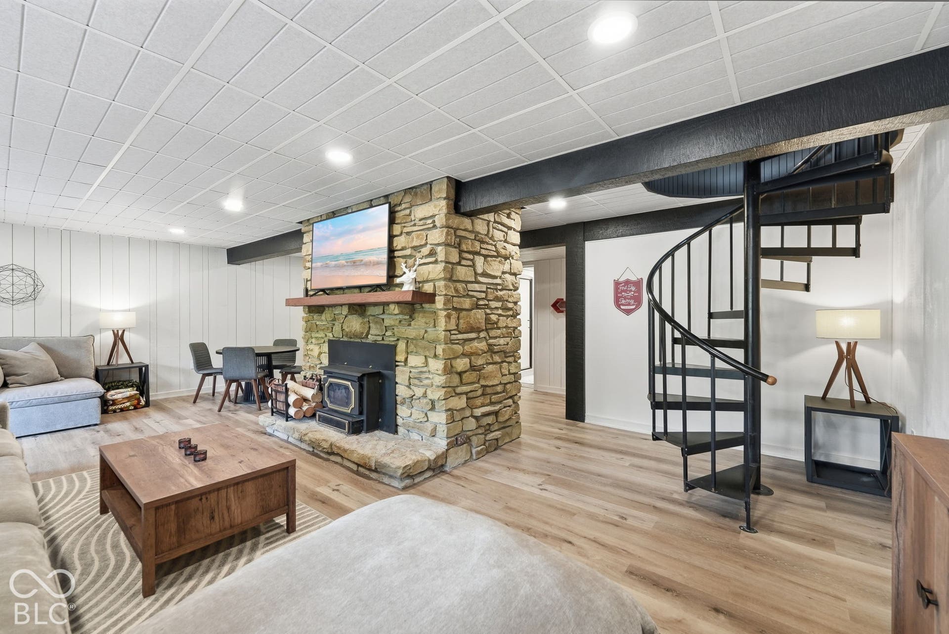 Stone-clad fireplace with wood-burning insert, wood-finish flooring, white vertical plank wall paneling, and a black spiral staircase
