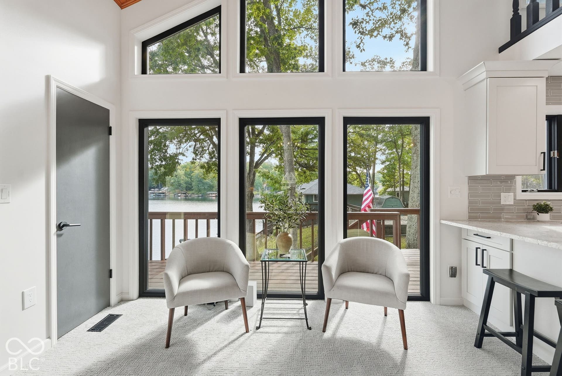 Vaulted ceiling with wood-finish planks, expansive window wall, light-toned carpeting, white cabinetry with dark hardware, and a light-toned countertop with a tile backsplash