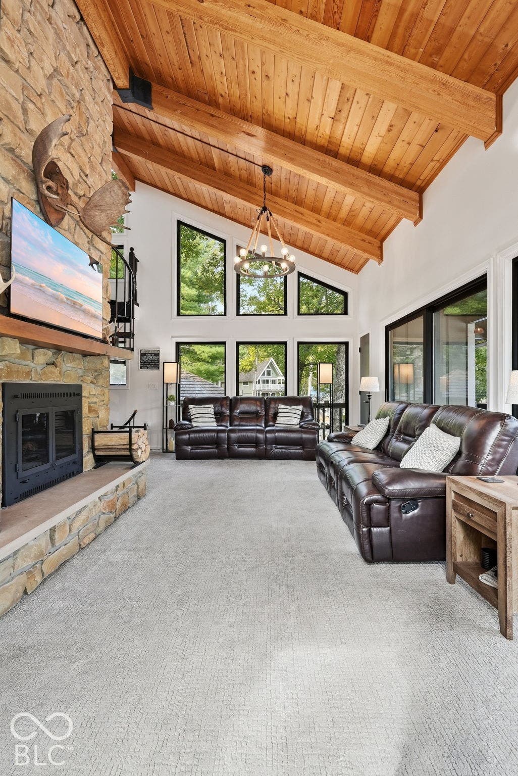 Vaulted wood plank ceiling with exposed beams, stone-clad fireplace, large picture windows, and light neutral carpeting