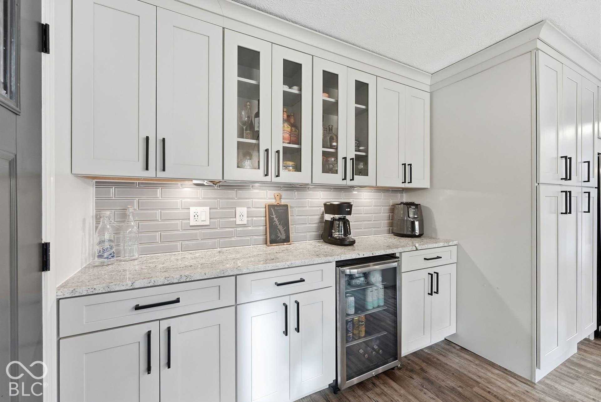 Bright, modern kitchenette featuring white shaker-style cabinetry with glass-front upper cabinets, matte black hardware, and a subway tile backsplash
