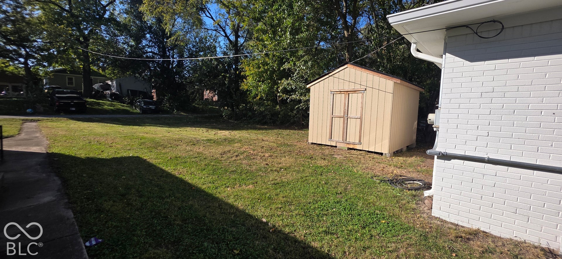 Expansive grassy yard featuring a detached storage shed with double doors, mature trees, and a concrete side path
