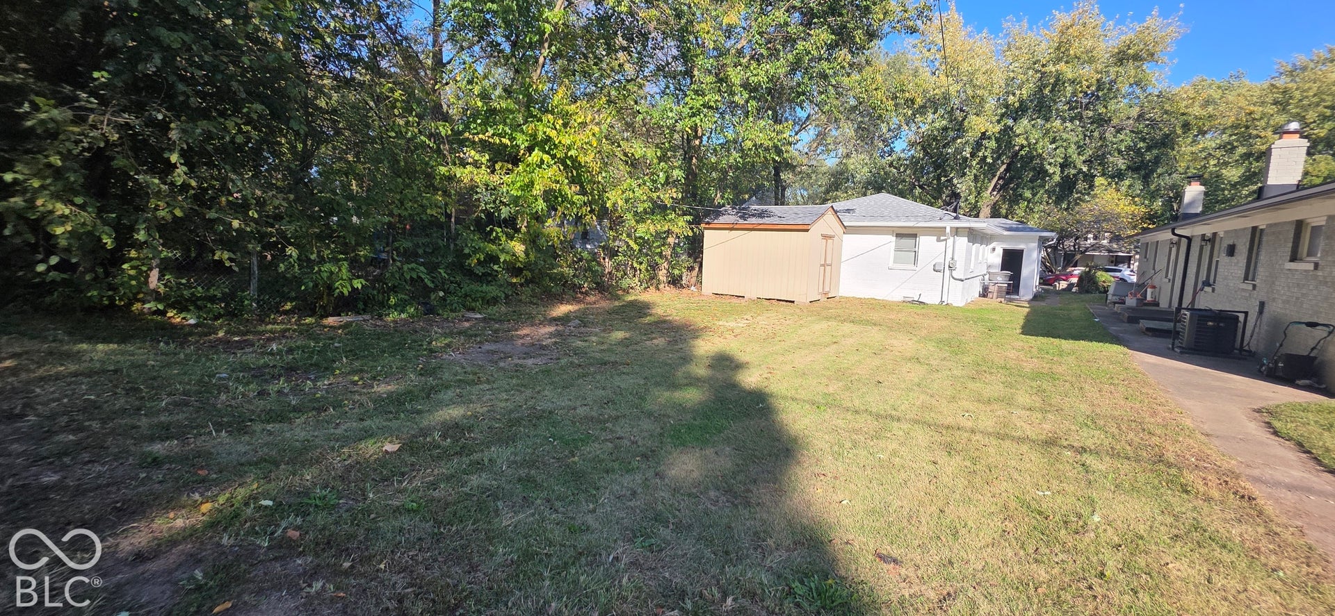 Expansive grassy area with mature perimeter trees, featuring a detached shed with a metal roof and a separate structure with white siding