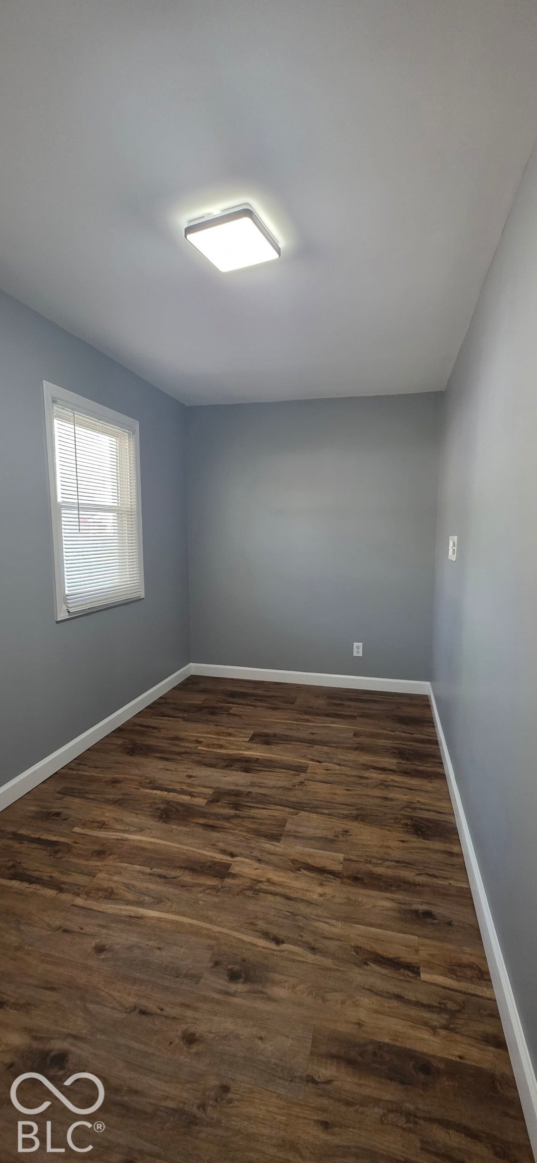 Wood-finish flooring with white trim, a single window, and a square surface-mount light fixture