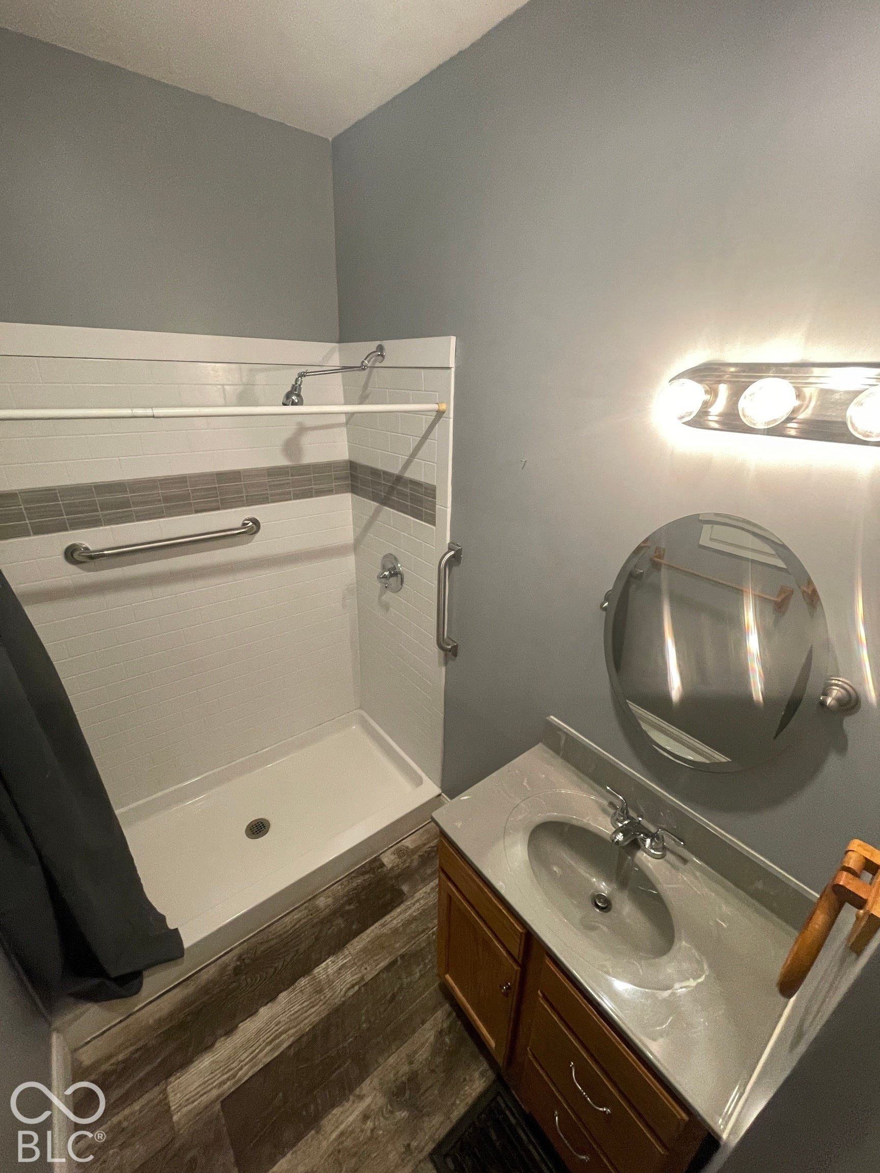 Walk-in shower with grab bars and tile surround, wood-finish flooring, vanity with integrated sink, and wall-mounted vanity lighting