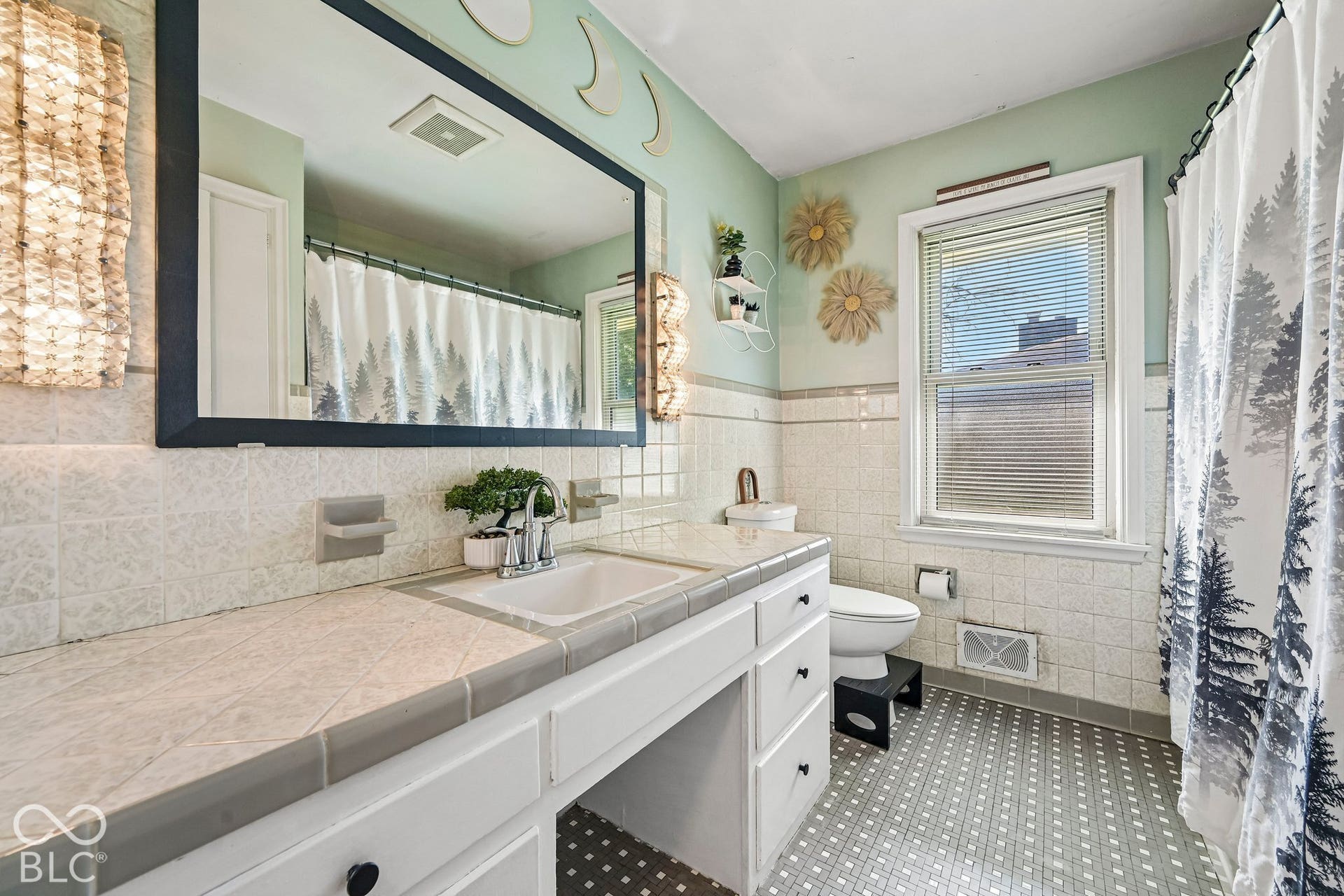Vanity with integrated sink, tile countertop, white cabinetry with dark hardware, large wall-mounted mirror, and tiled flooring