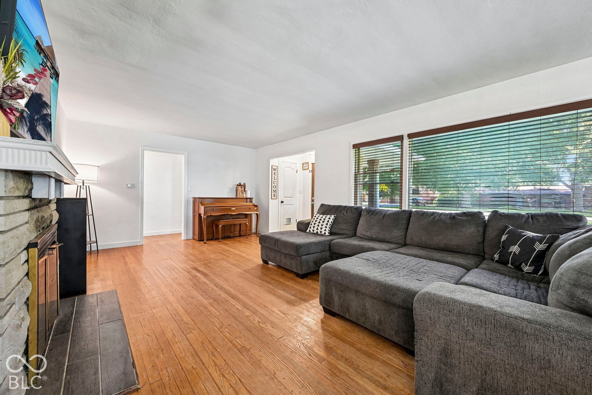 Hardwood flooring, white walls, stone-faced fireplace with mantel, and large windows with horizontal blinds