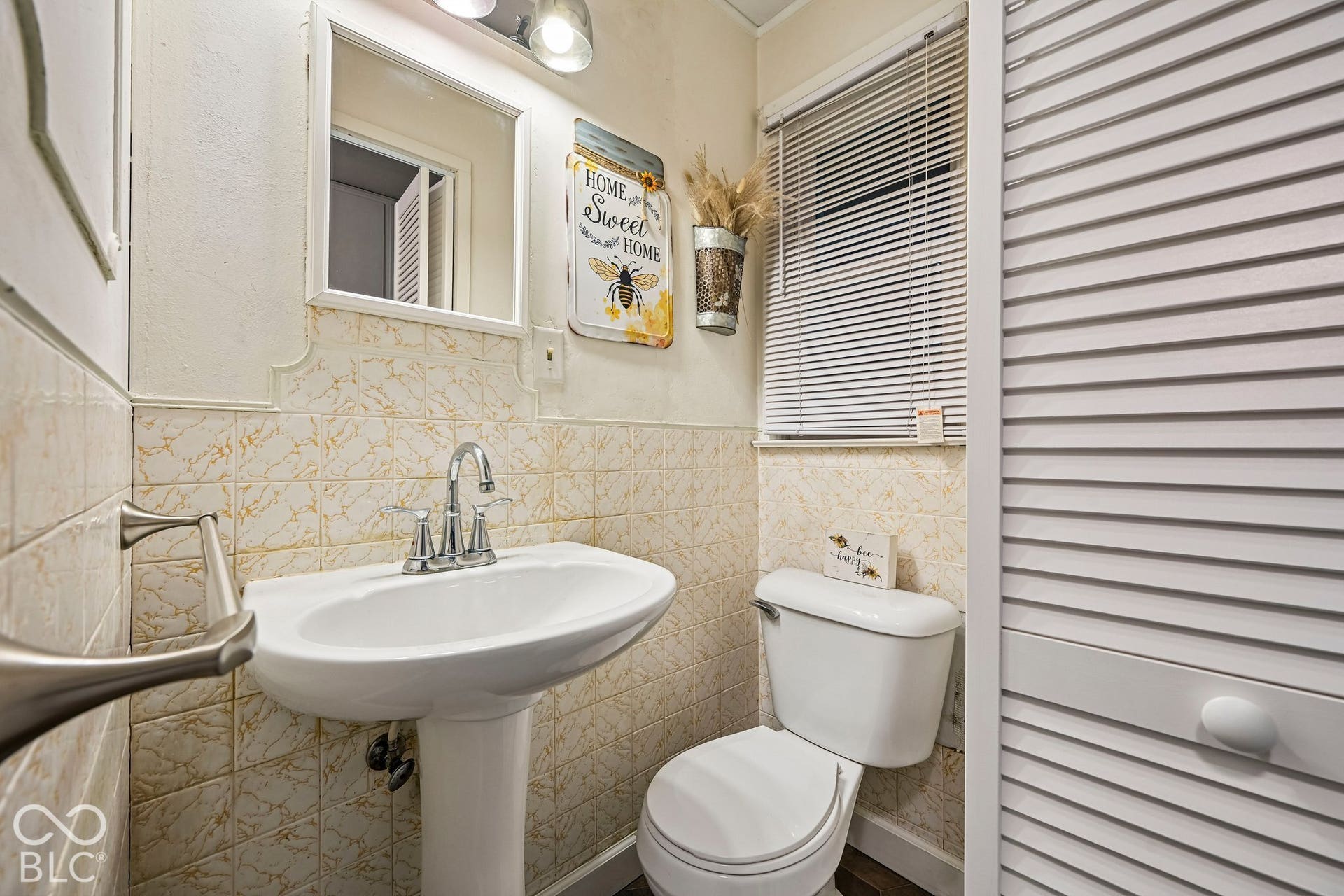 Pedestal sink with chrome faucet, wall-mounted mirror, ceramic tile wainscoting, toilet, and louvered bifold doors