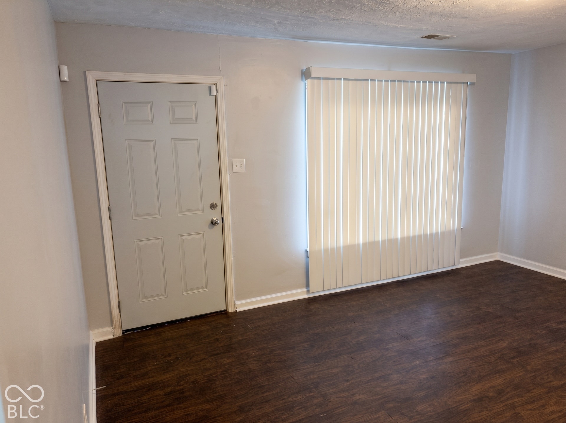 Dark wood-finish flooring, light grey walls, white trim, paneled door, and vertical blinds
