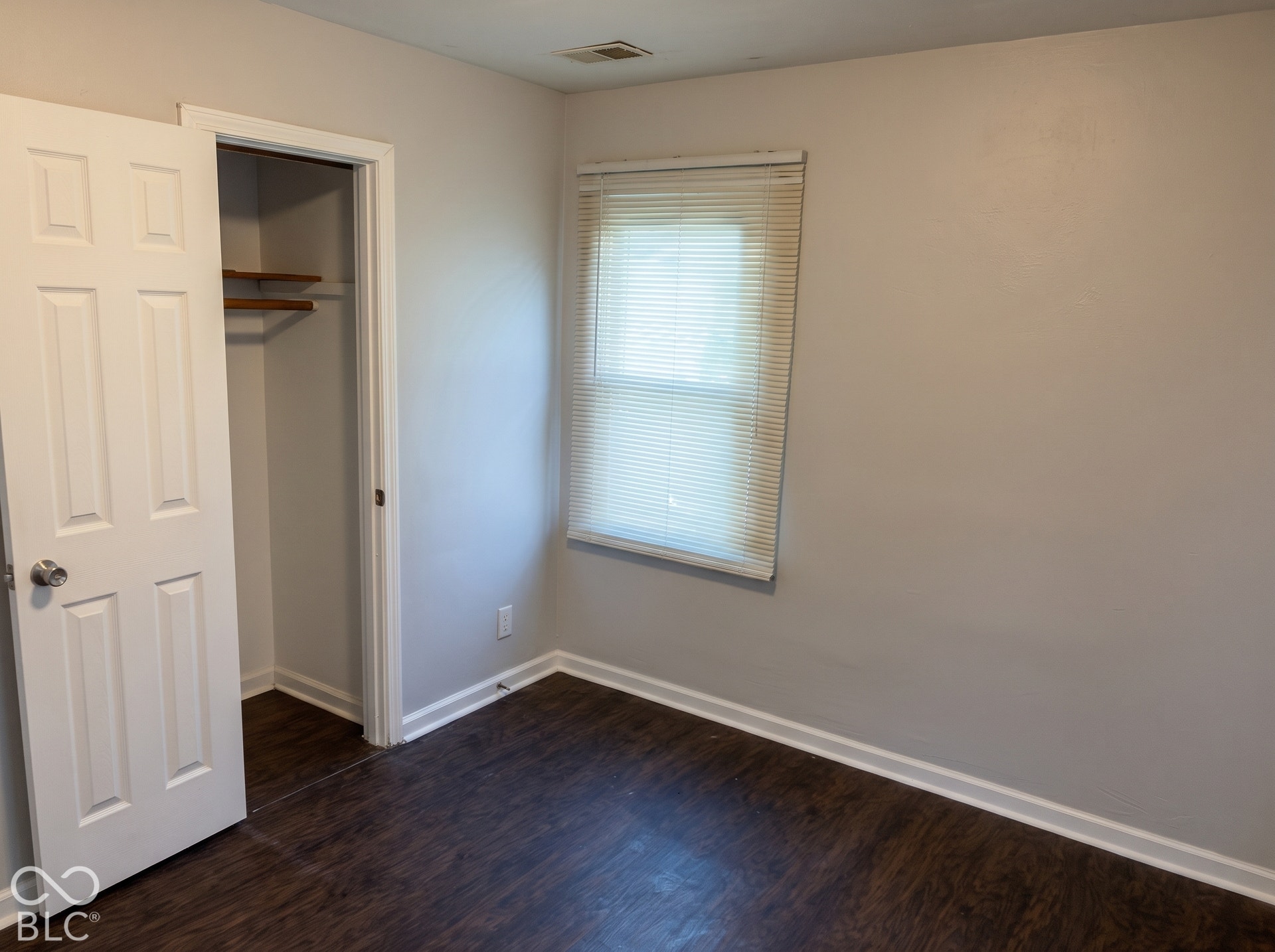 Wood-finish flooring with white trim, a single window with blinds, and light-toned walls