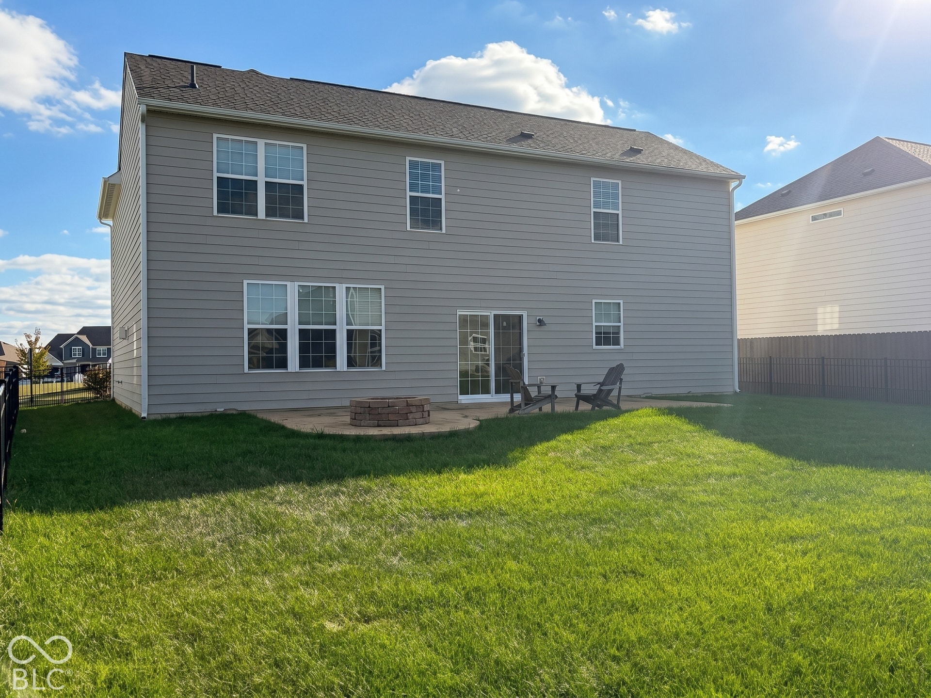 Virtually Altered - Expansive lawn with a black metal fence, concrete patio, and a built-in fire pit