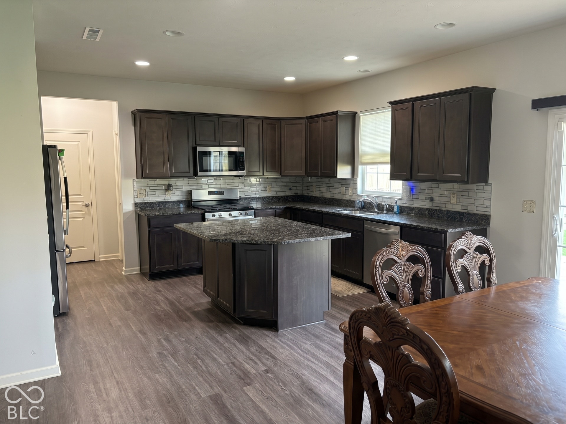 Virtually Altered - Kitchen featuring wood-finish flooring, dark wood cabinetry, a center island with granite-style countertop, a tile backsplash, and stainless steel appliances