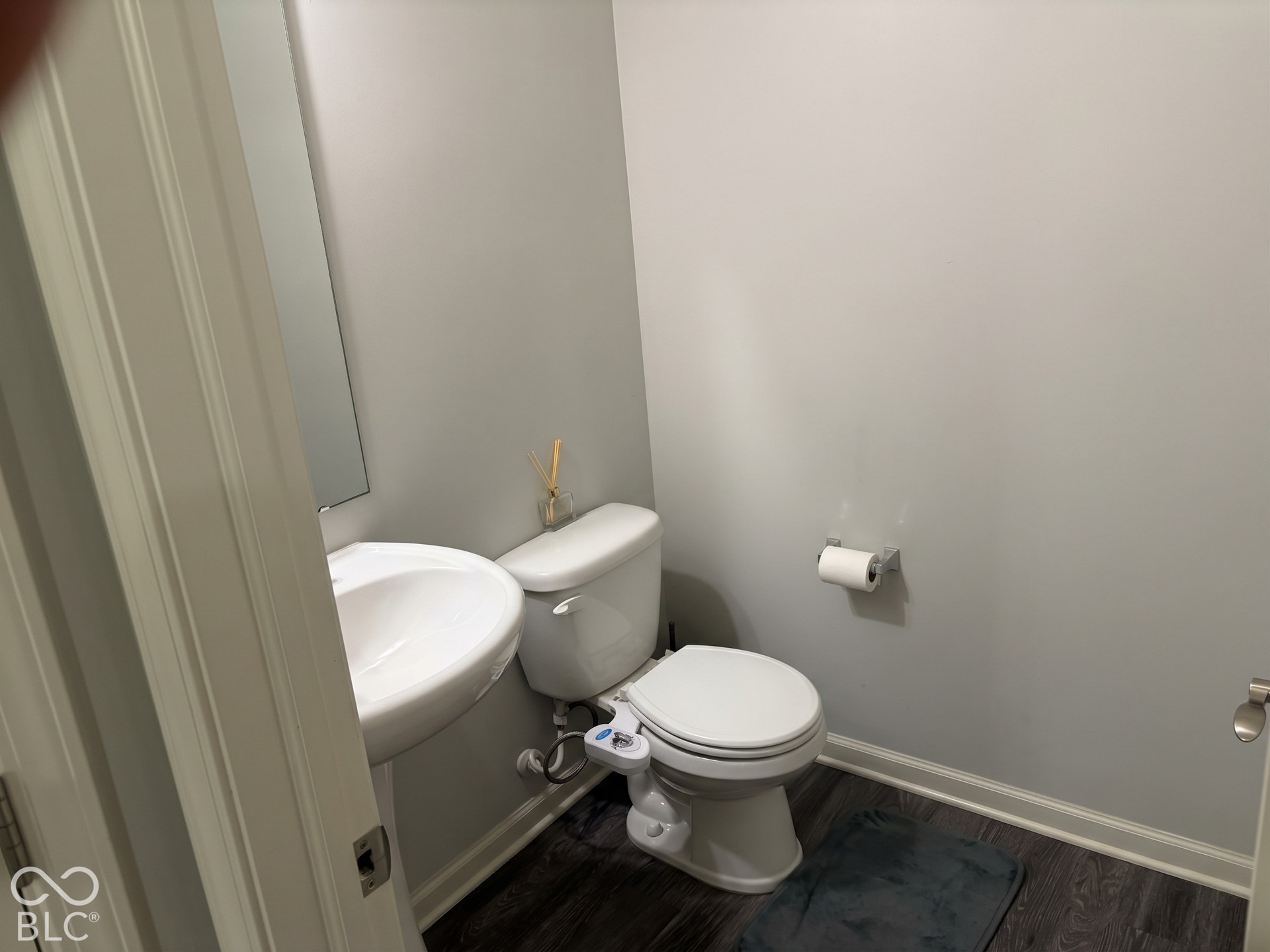 Virtually Altered - Main-level half bath with pedestal sink with a wall-mounted mirror, white toilet, and wood-finish flooring