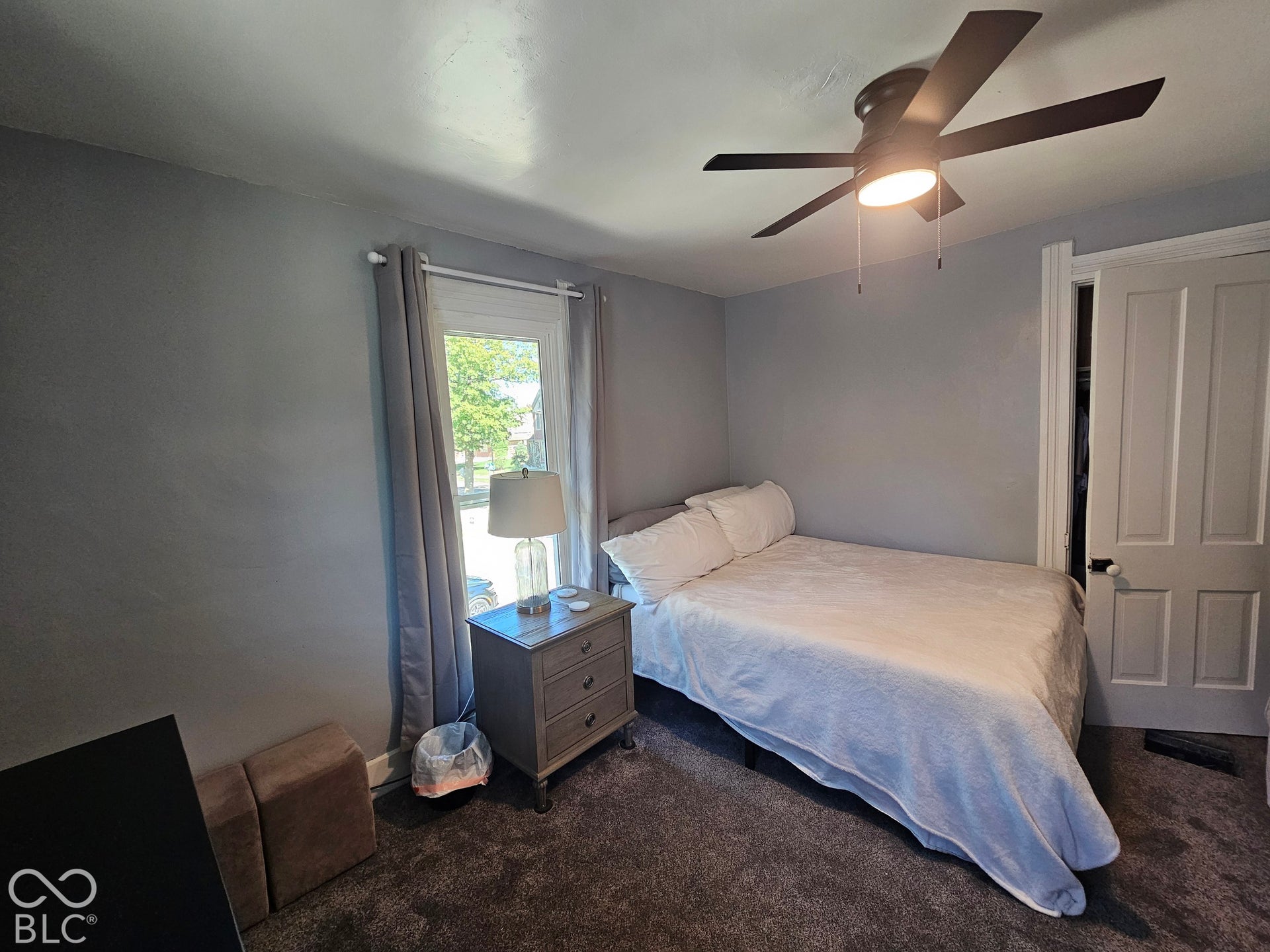 917 W 8th Street, Anderson, IN 46016 Dark-toned wall-to-wall carpeting, window with light-toned curtains, gray painted walls, and a ceiling fan with integrated lighting