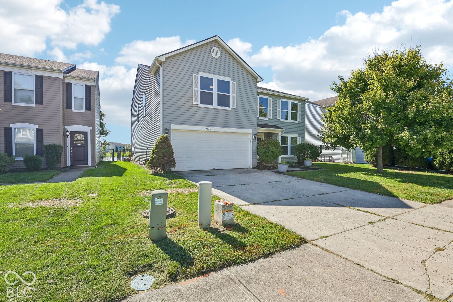 Two-story exterior with light blue siding and white window shutters, attached garage, concrete driveway, and mature landscaping