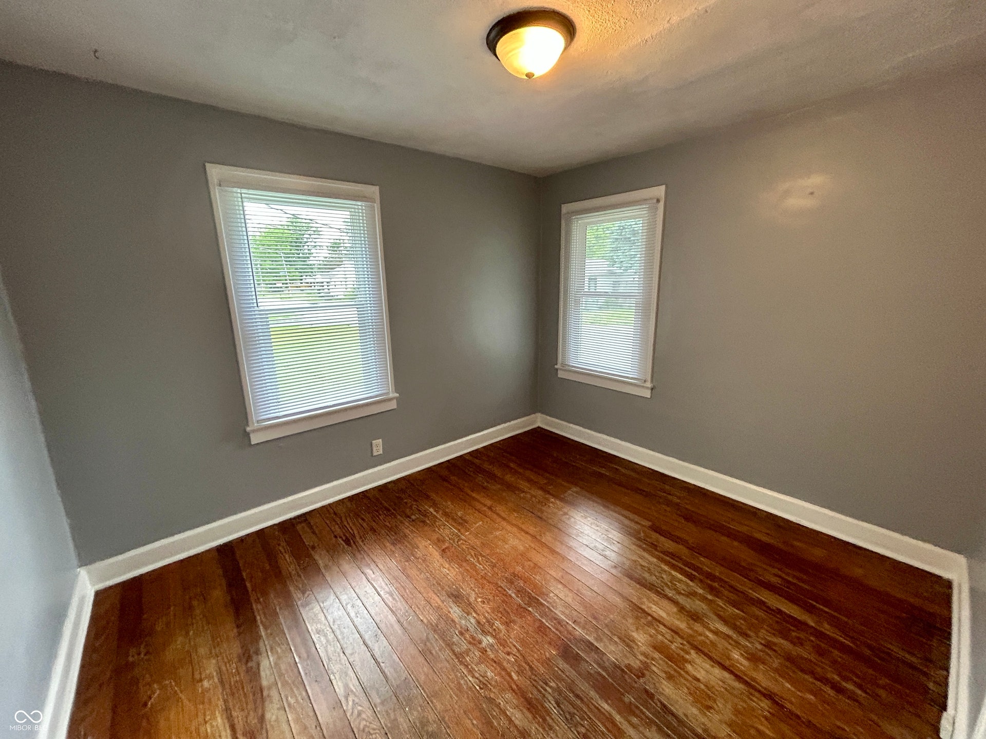 Hardwood flooring throughout, two windows with white blinds, neutral gray wall tones, and a flush-mount ceiling light fixture