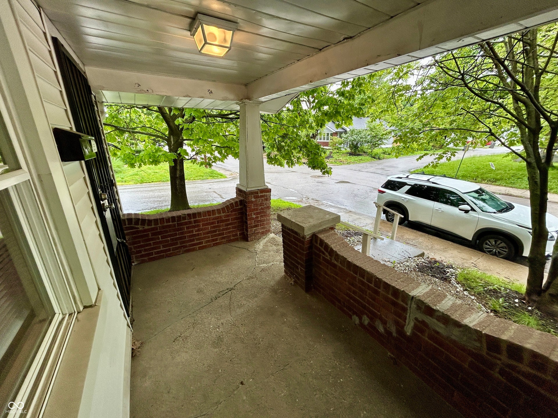 Covered front porch featuring a brick half-wall, concrete flooring, and a ceiling-mounted light fixture