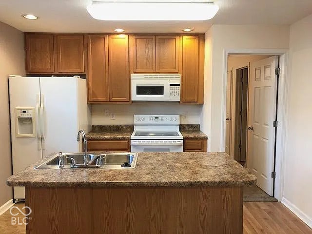 Kitchen featuring wood cabinetry, an island with a double basin sink, white appliances, and wood-finish flooring