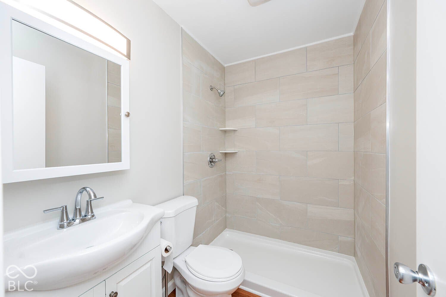White vanity with chrome faucet and integrated sink, mirrored medicine cabinet with overhead lighting, tiled shower surround with corner shelves, and a white shower pan