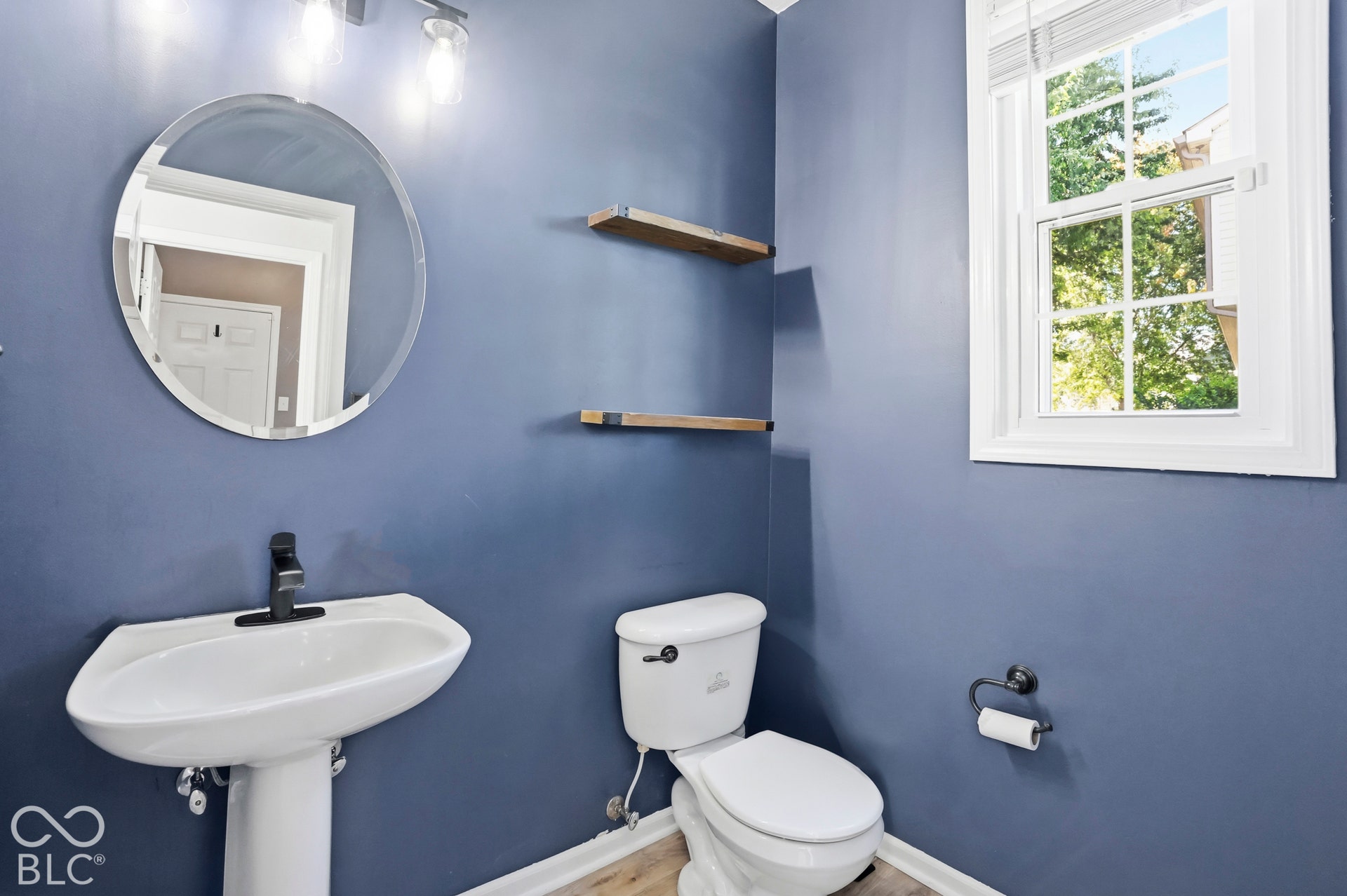 Pedestal sink with a black-finish faucet, circular wall-mounted mirror, and decorative wall shelving
