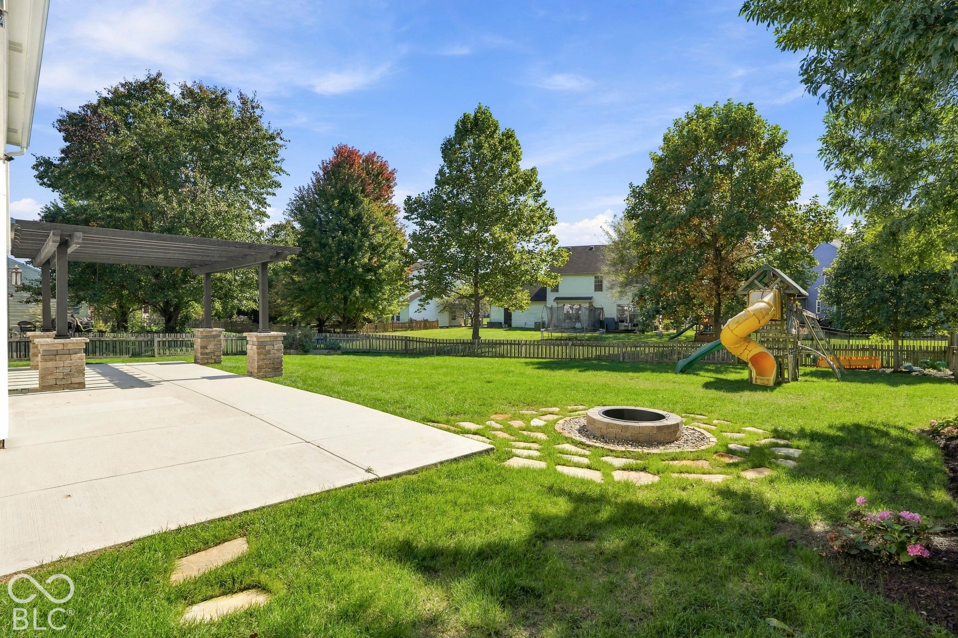 Expansive backyard featuring a concrete patio, a dark wood pergola with stone-clad columns, a circular fire pit with stepping stones, and a manicured lawn