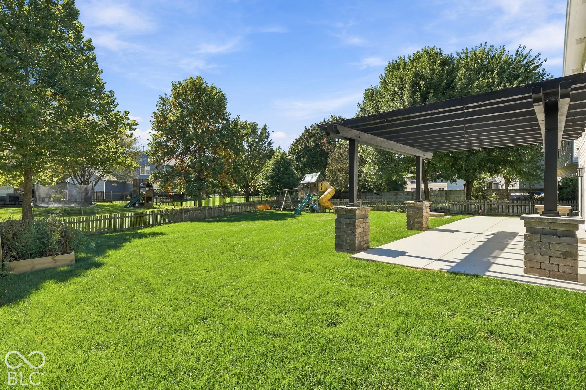 Expansive manicured lawn with a privacy fence, mature trees, and a concrete patio featuring a dark-toned pergola with stone-clad pillars