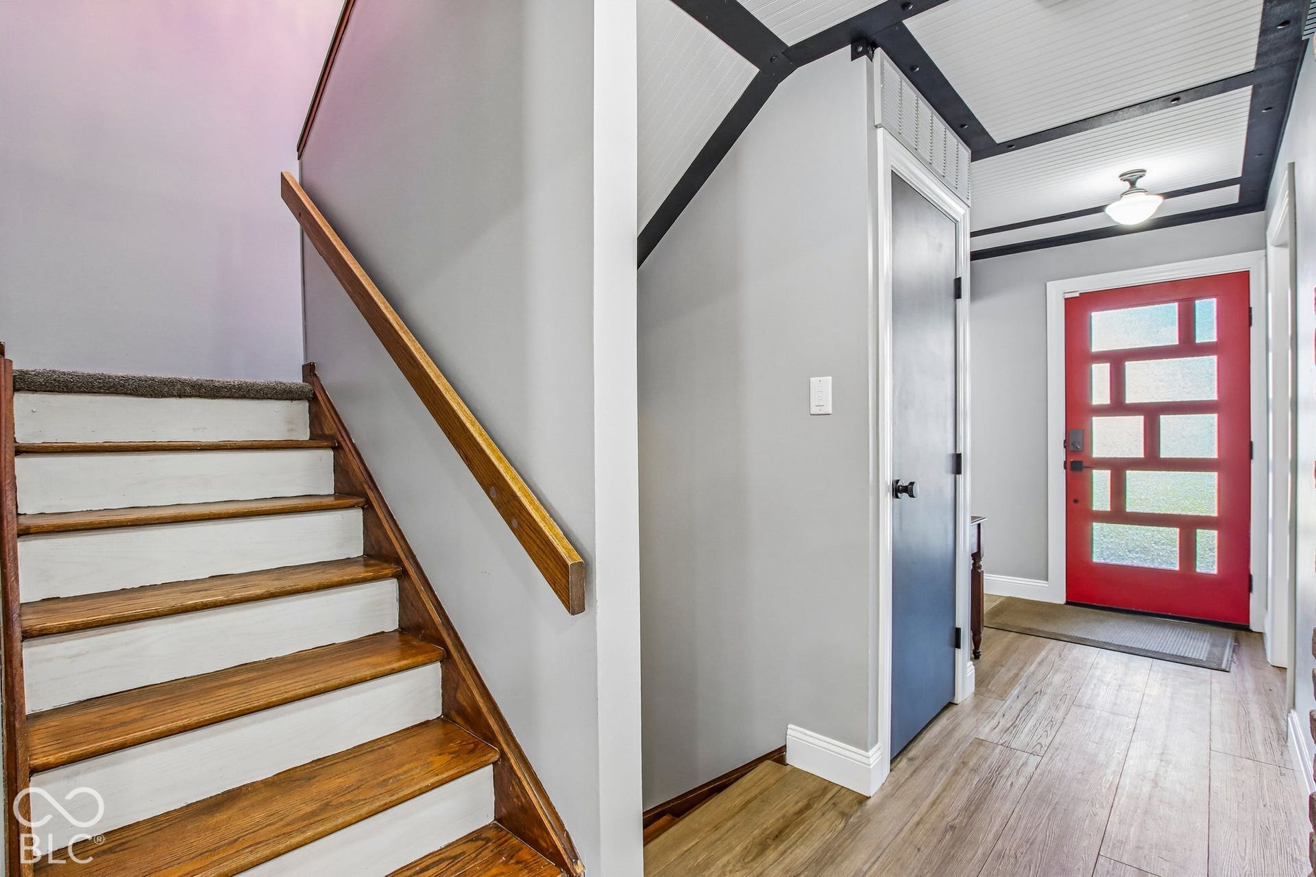 Wood-finish flooring, staircase with wood treads and handrail, red entry door with multiple glass panels, black trim ceiling with recessed panels, and flush-mount ceiling light fixture