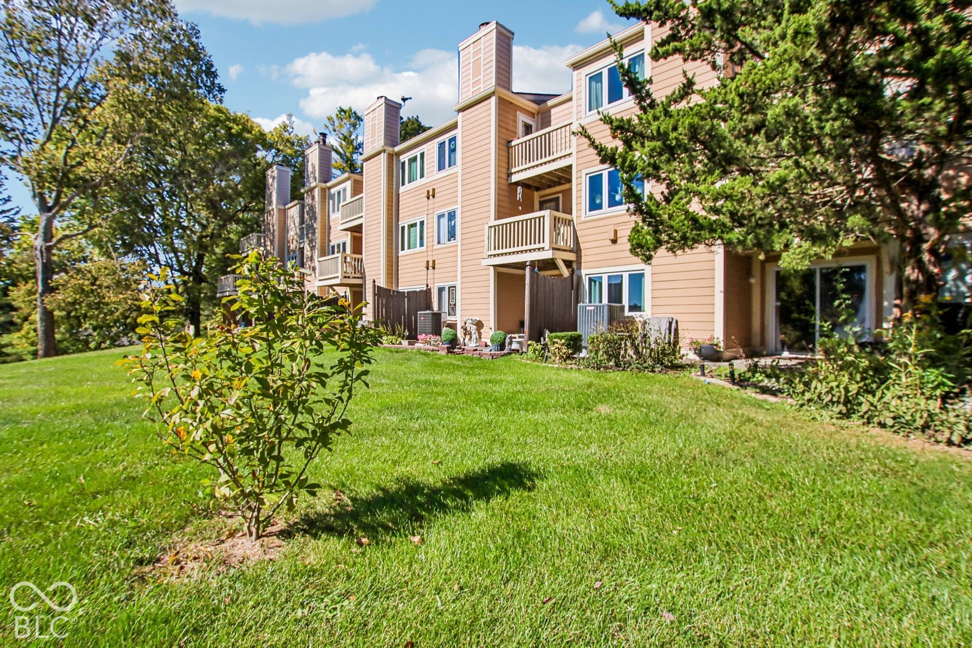 Expansive manicured lawn with mature landscaping and newly planted specimen trees, backing a multi-story residence featuring wood-finish siding, blue-trimmed windows, and multiple balconies with railings