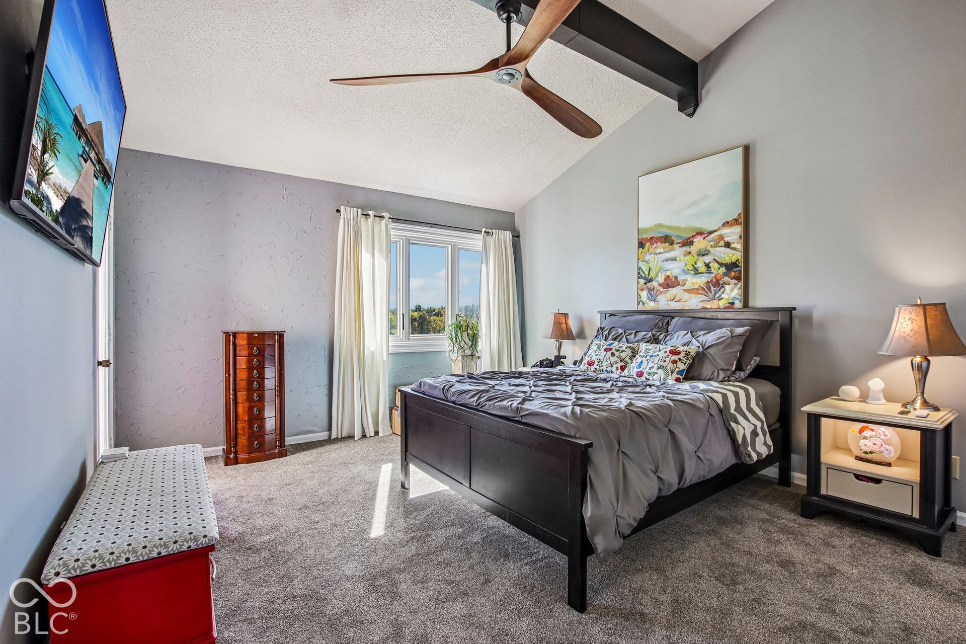 Vaulted ceilings with exposed beams, wood-finish ceiling fan, large window with natural light, and gray carpeting