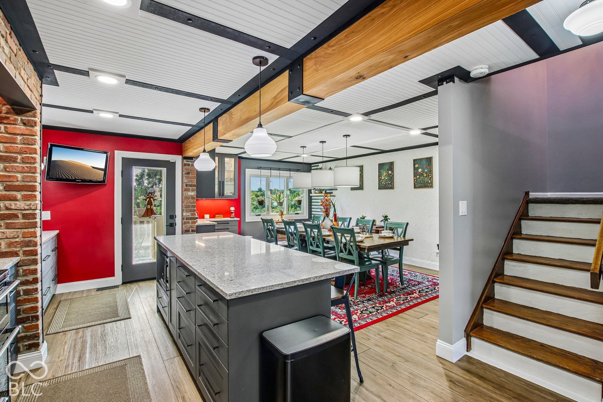 Exposed brick accent wall, wood-finish flooring, kitchen island with granite countertop and built-in cabinetry, exposed wood ceiling beams, and a dining area with pendant lighting