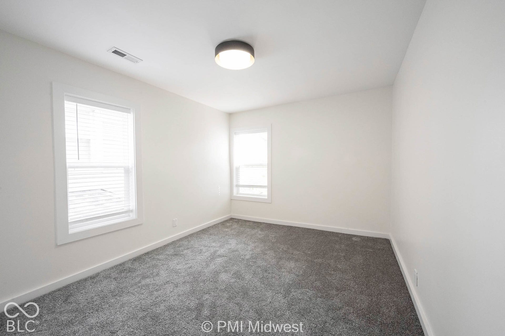 Neutral white walls with gray carpeting, two windows with white blinds, and a flush-mount ceiling fixture