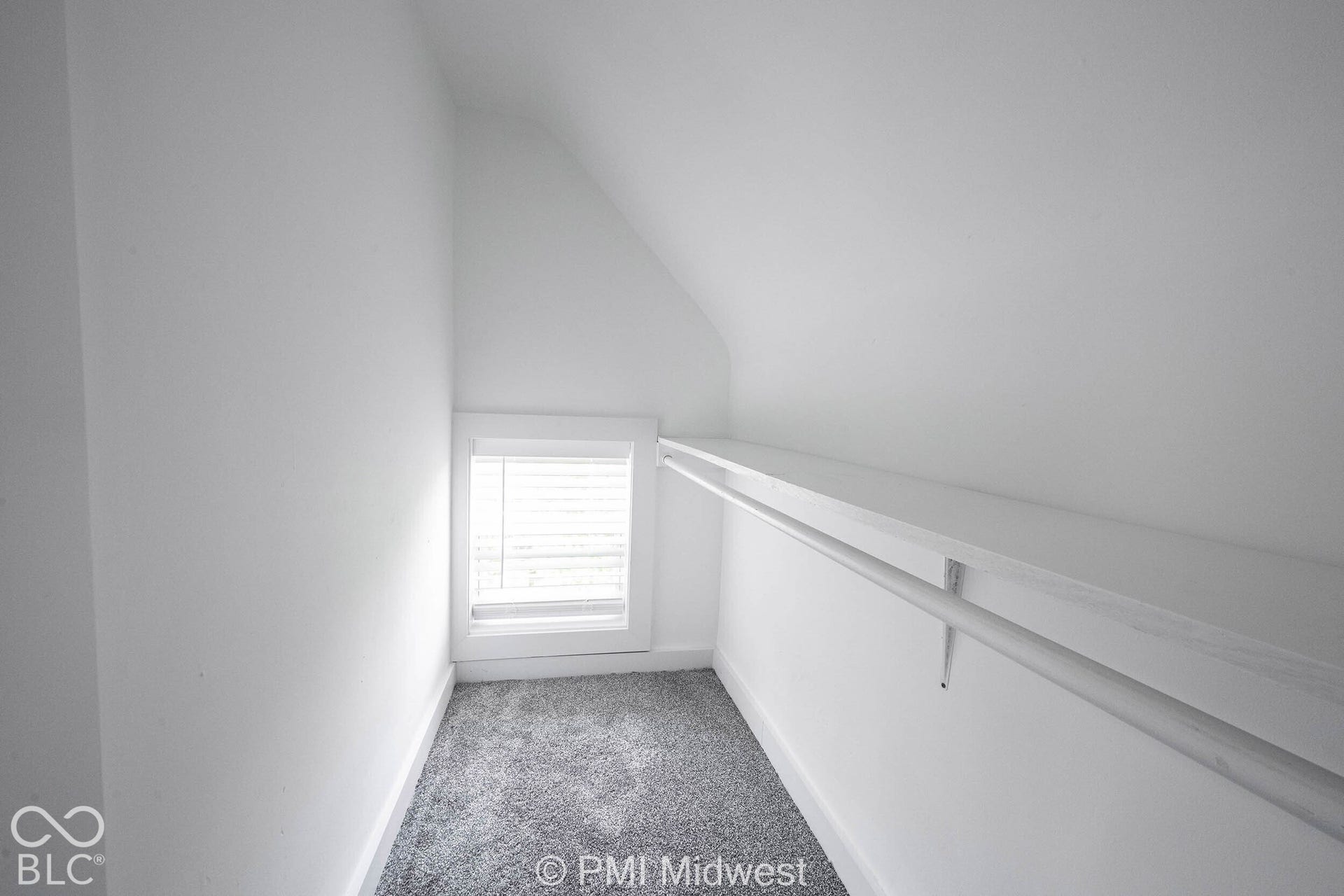 Attic space with sloped ceilings, a single window with blinds, and built-in shelving with a hanging rod